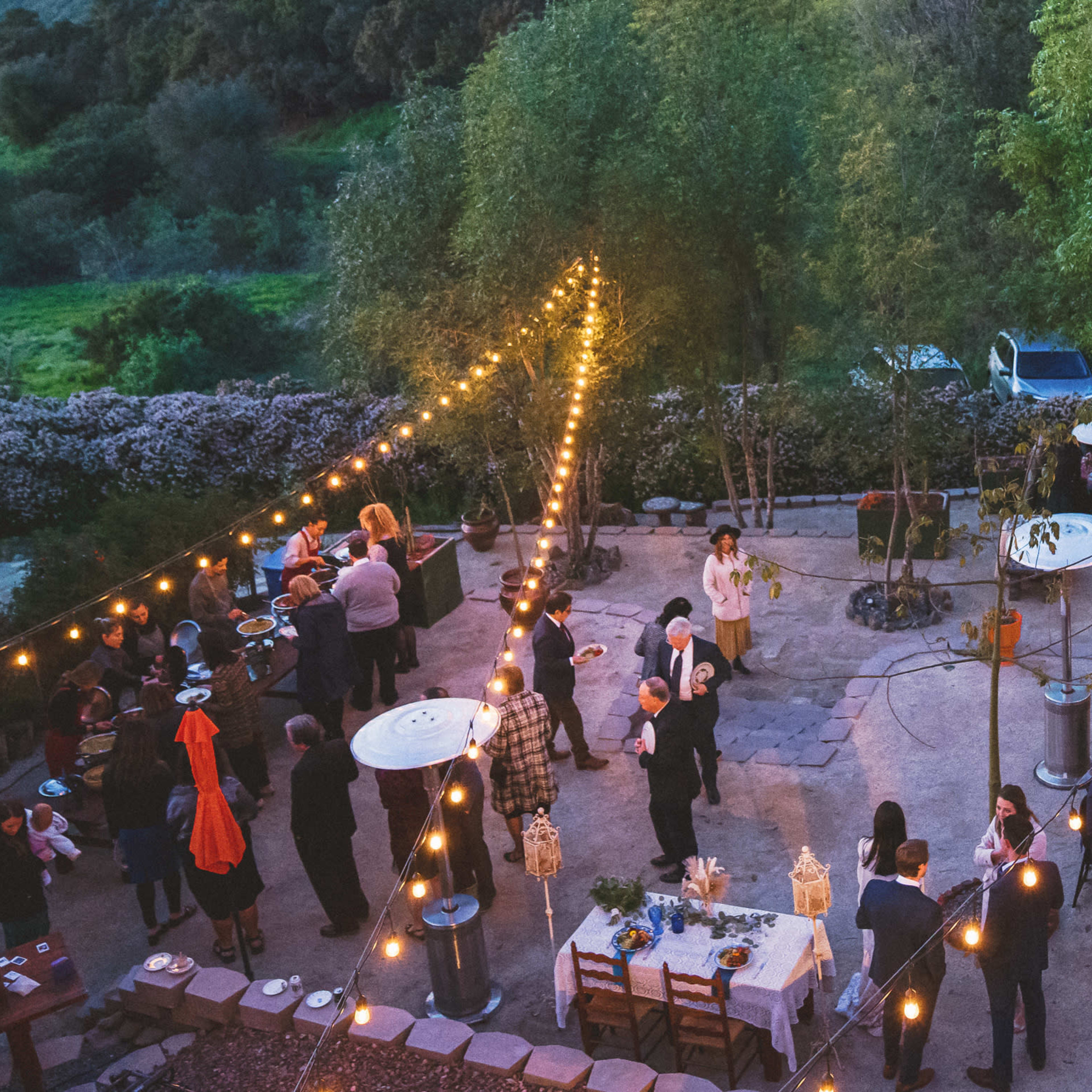 A gathering takes place outdoors in a landscaped area, illuminated by string lights, with guests mingling around tables set for a meal.