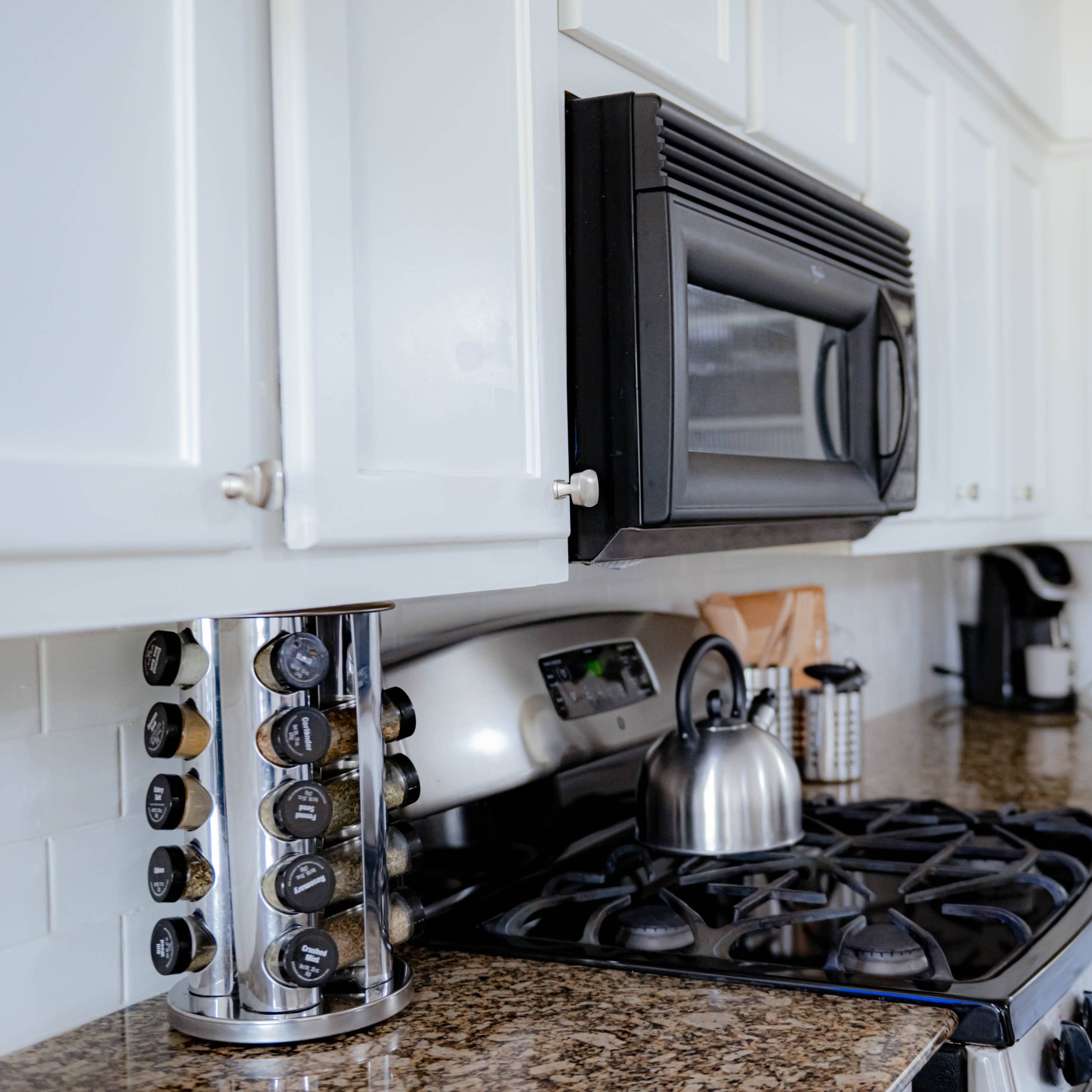 A modern kitchen with white cabinets, a black microwave, a kettle on the stove, and a spice rack on the countertop.