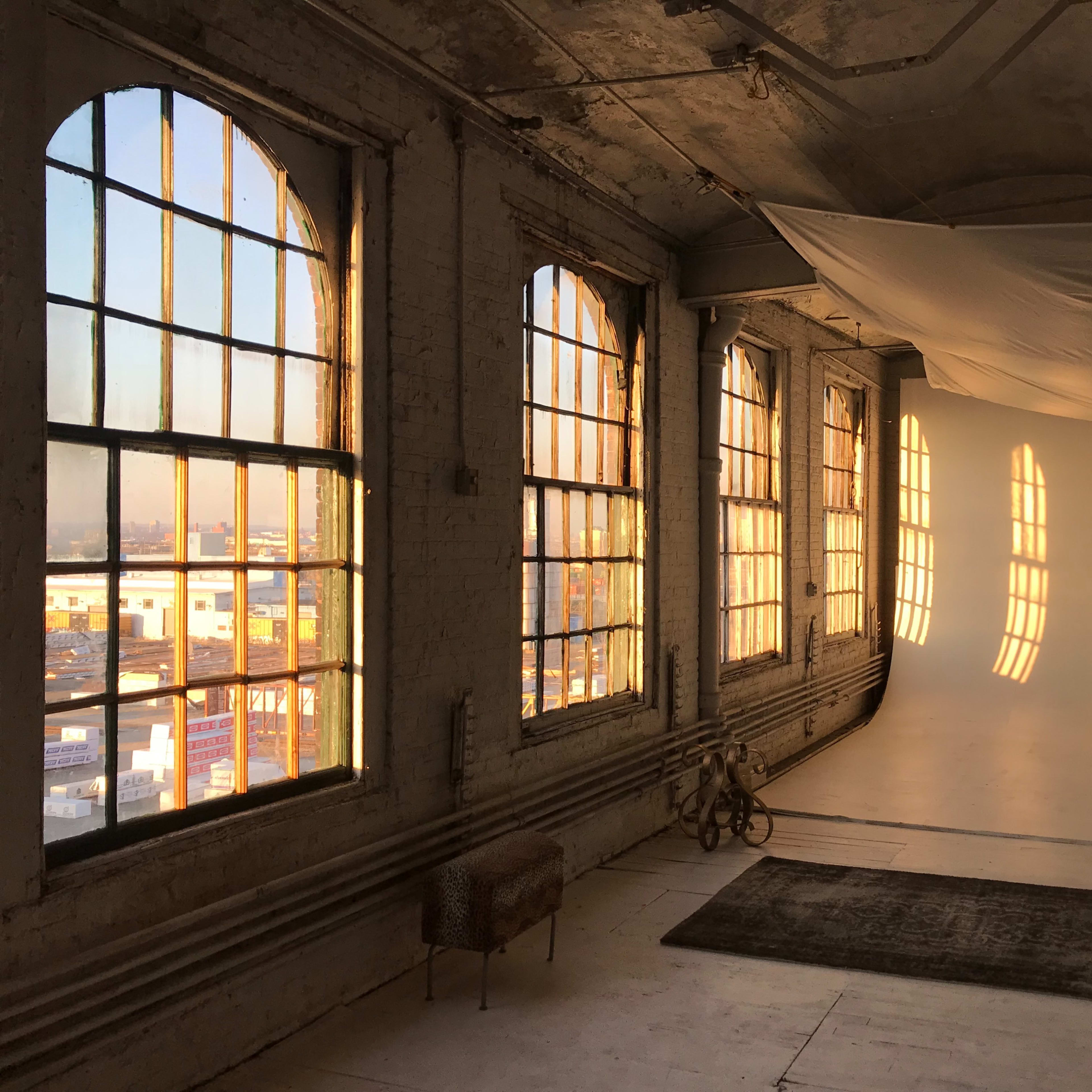A spacious studio with large, arched windows allowing sunlight to cast shadows on a white backdrop and wooden floor.