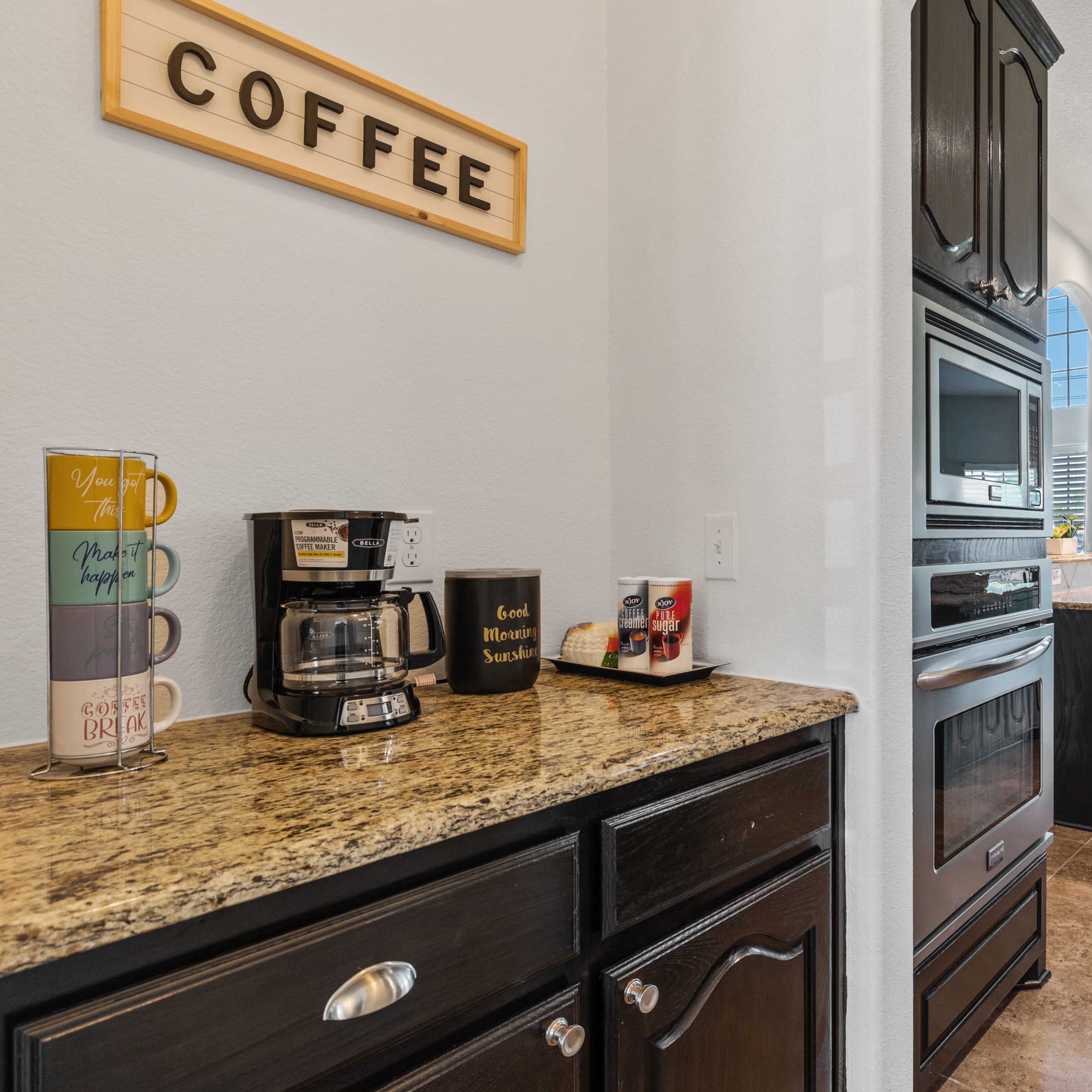 A well-lit kitchen features a coffee maker on a granite countertop, with dark cabinetry and appliances, opening to a dining area through a doorway.