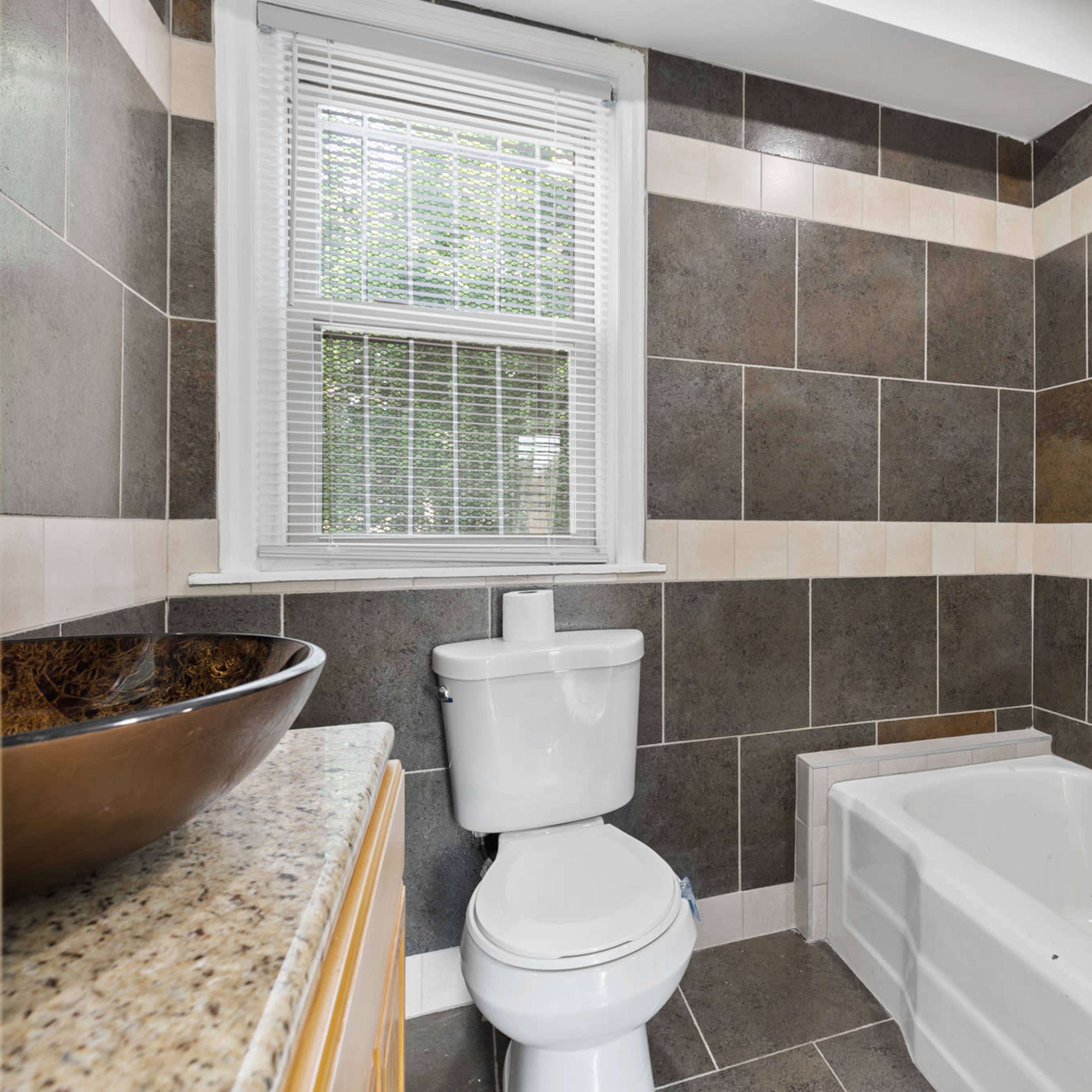 A bathroom featuring a white toilet, a bathtub, and slate-colored tiled walls with a marble countertop.