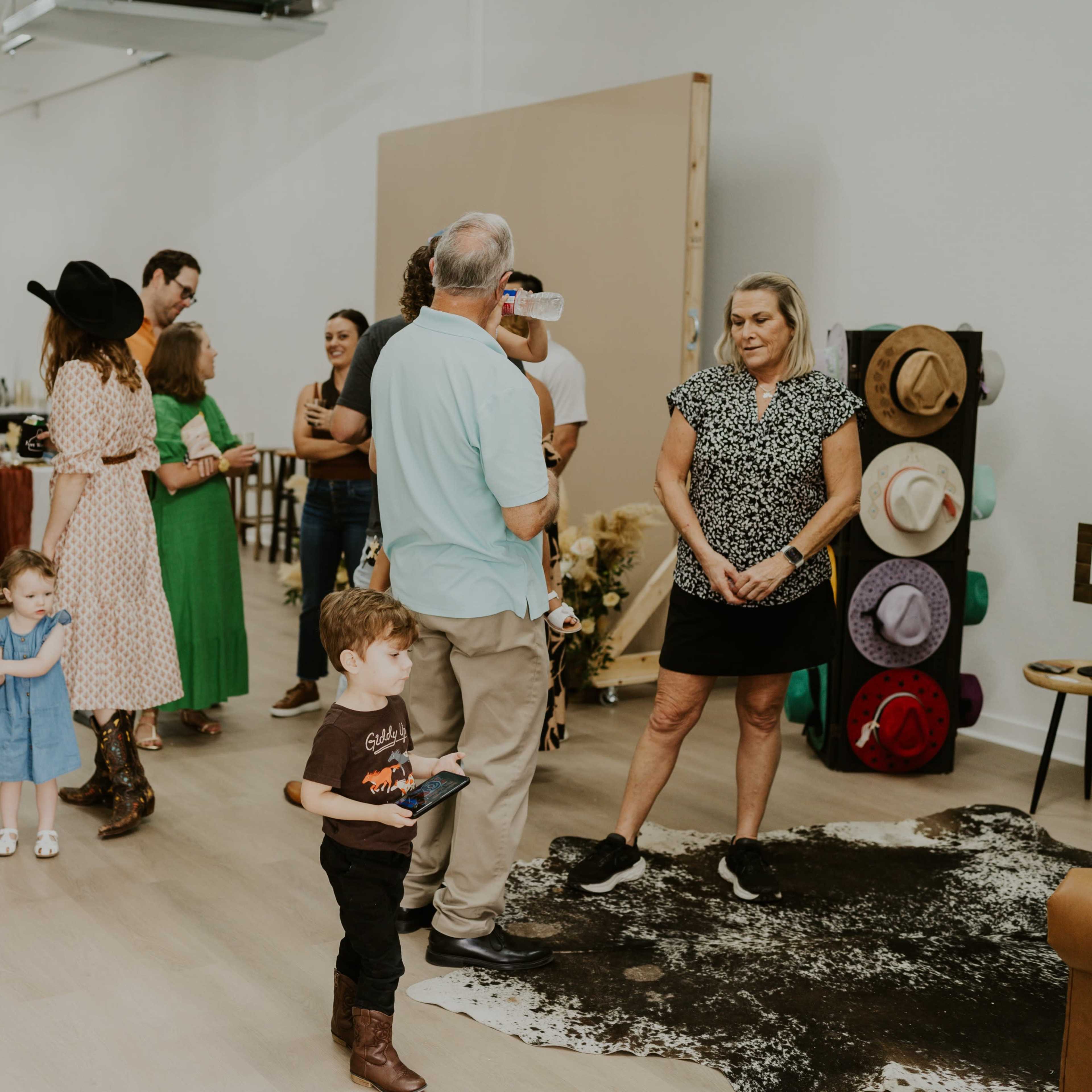 A diverse group of people gathers in a spacious studio, engaging in conversation and examining items displayed on tables, with various hats and decor in the background.