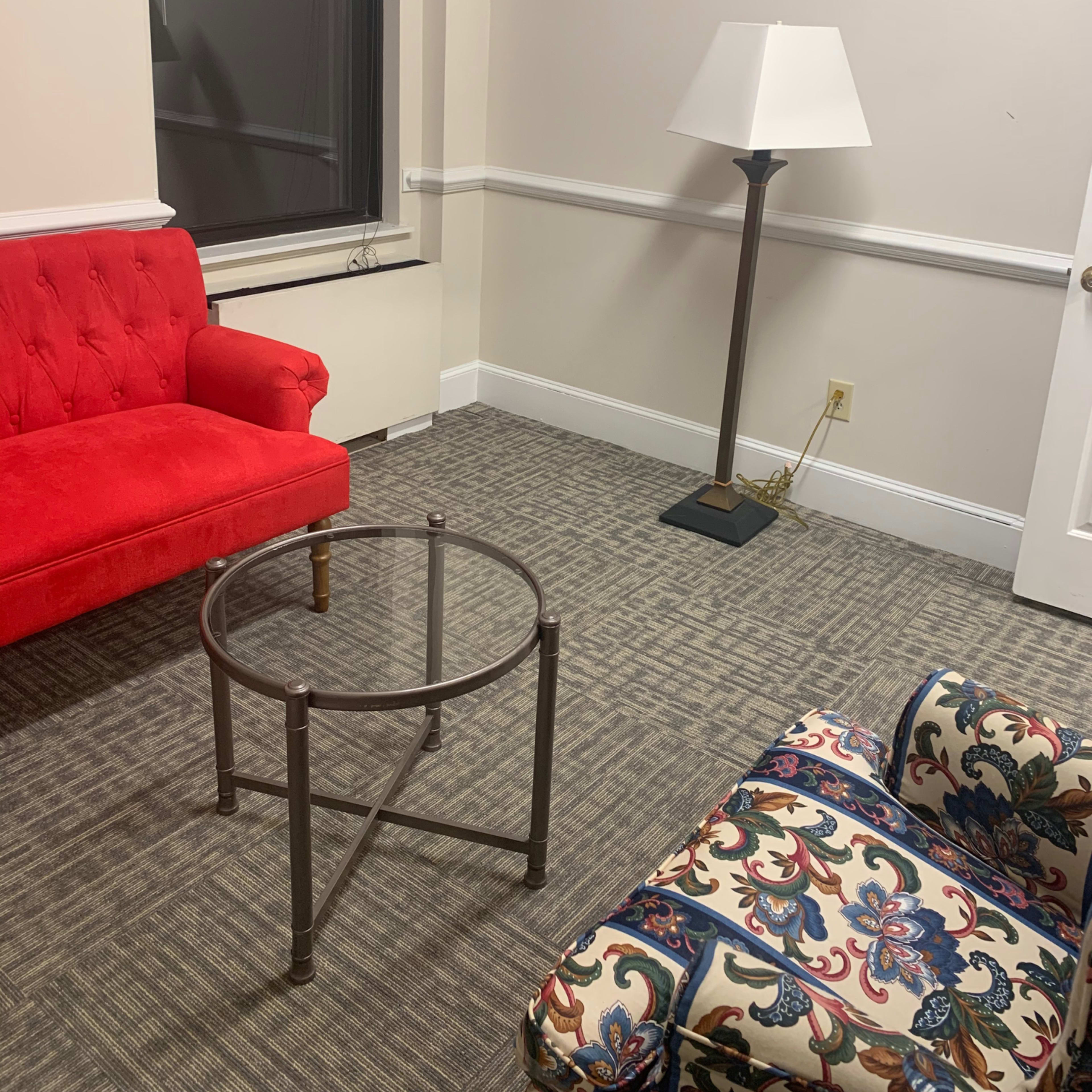 A small room with a red sofa, a patterned chair, a round glass table, and a floor lamp.