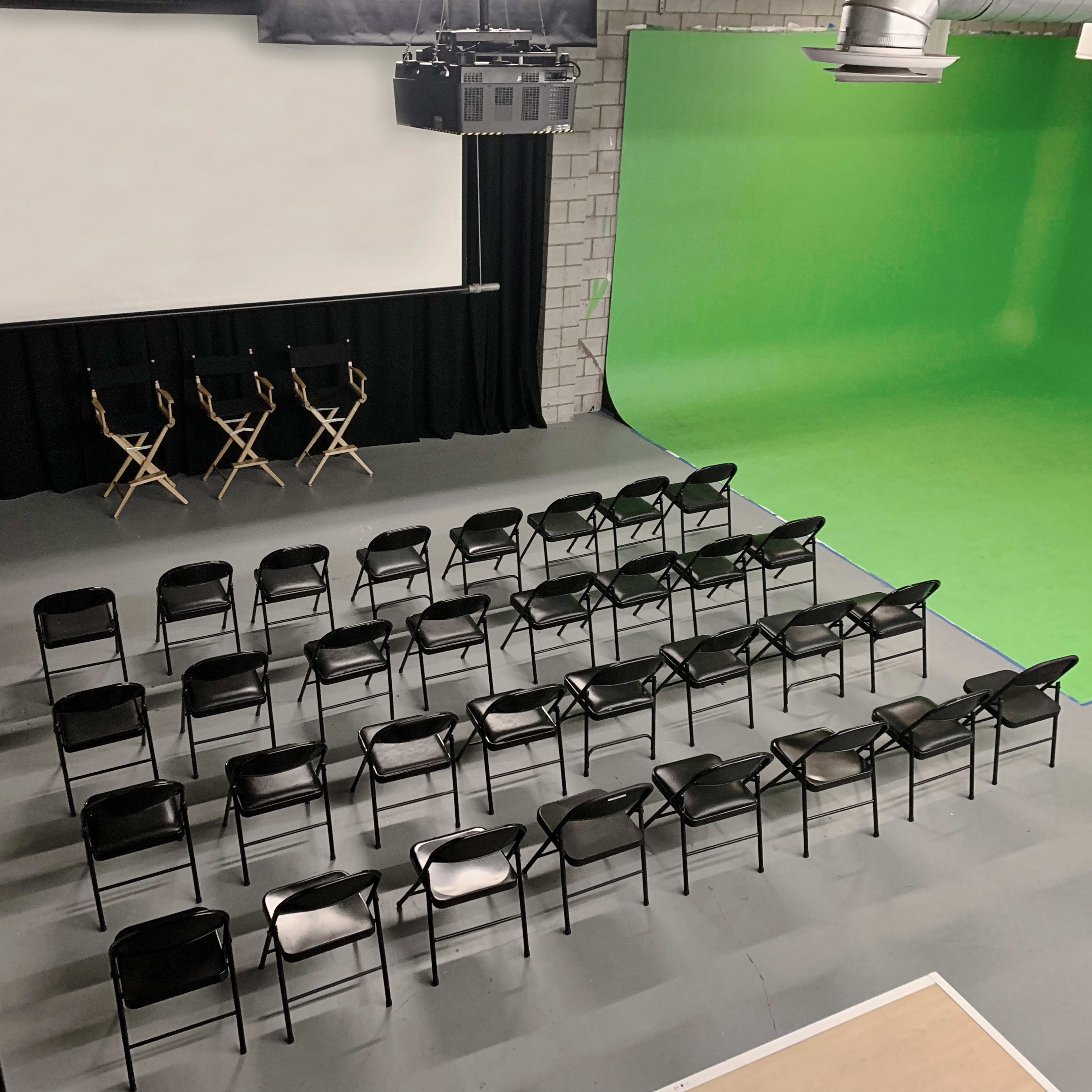 A film studio space with a green screen, three director's chairs, and rows of black chairs arranged in front.