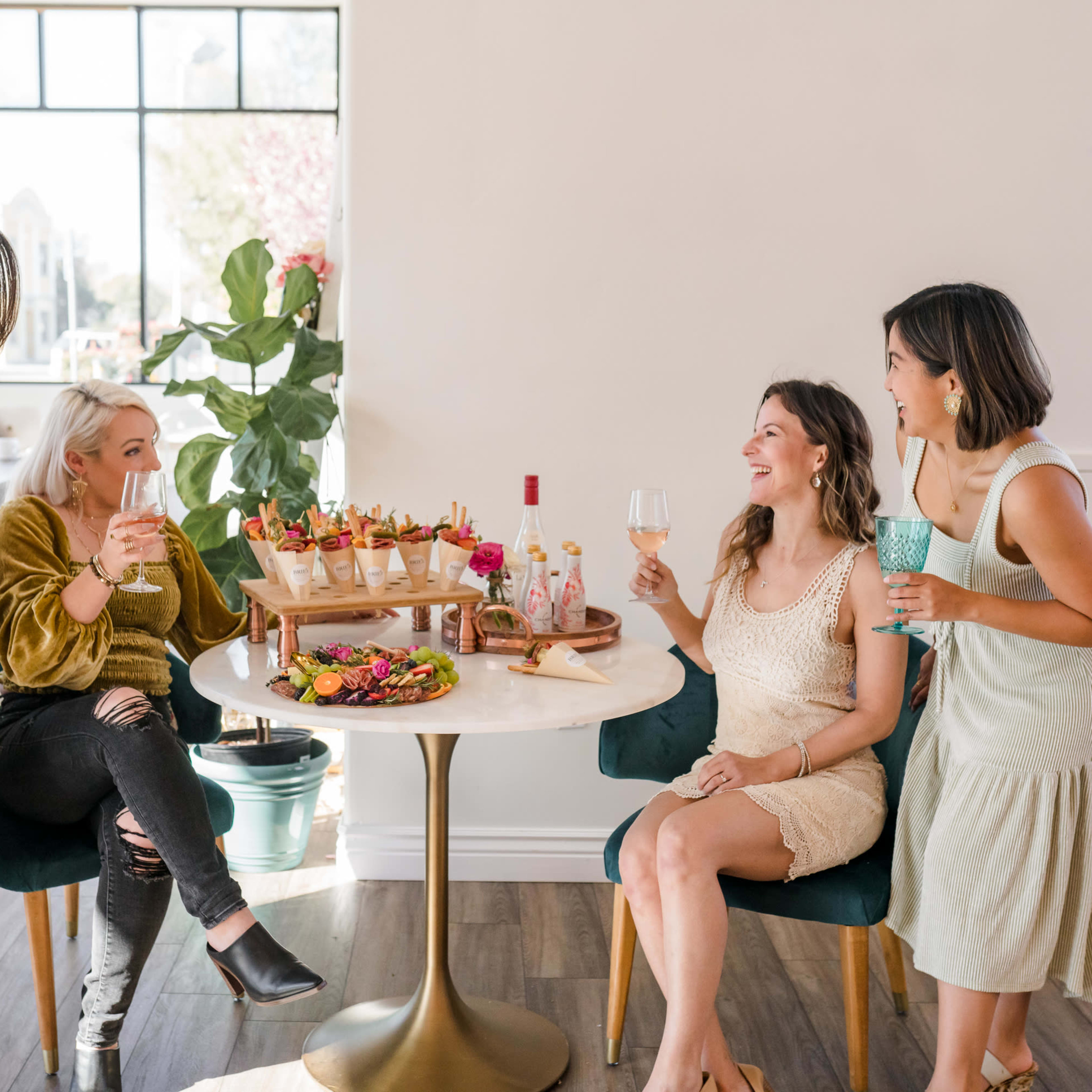 Four women are gathered around a table, enjoying colorful cocktails and a platter of food in a well-lit room with large windows.