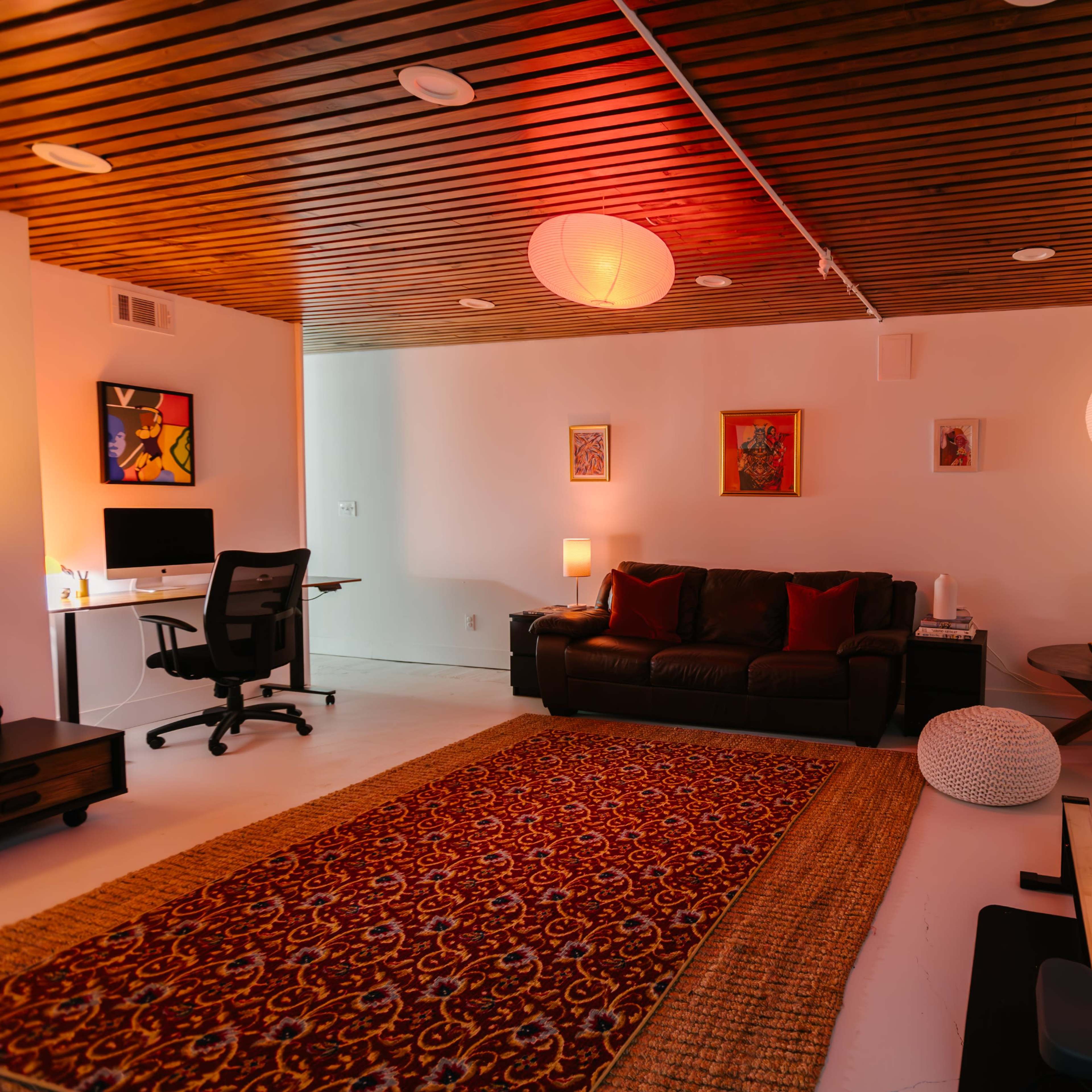 A spacious room featuring a brown couch, a wooden ceiling, a decorative rug, a desk with a computer, and soft lighting from paper lanterns.