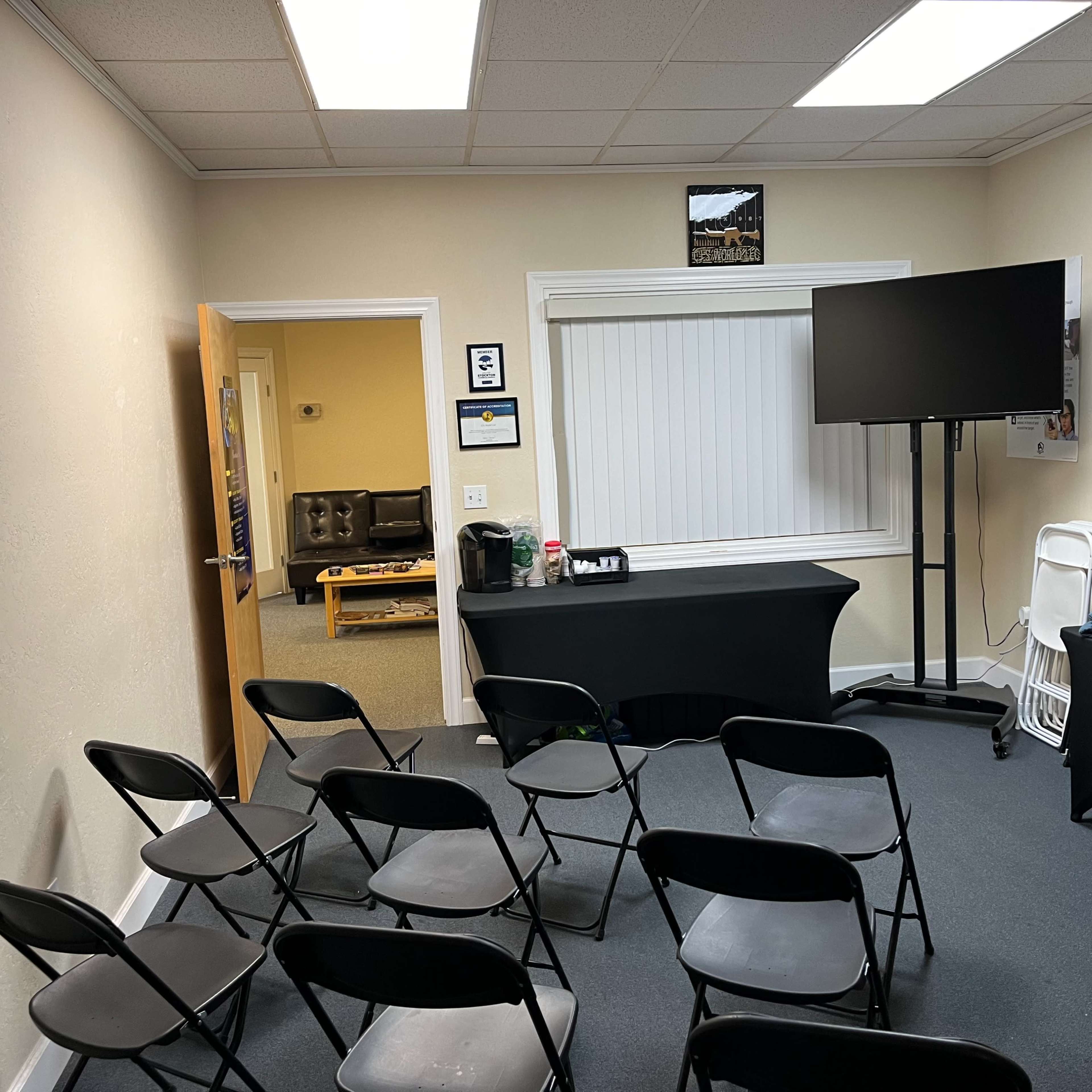A small meeting room with black folding chairs arranged in rows, a table covered with a black cloth, and a large screen on a stand opposite a window with blinds.
