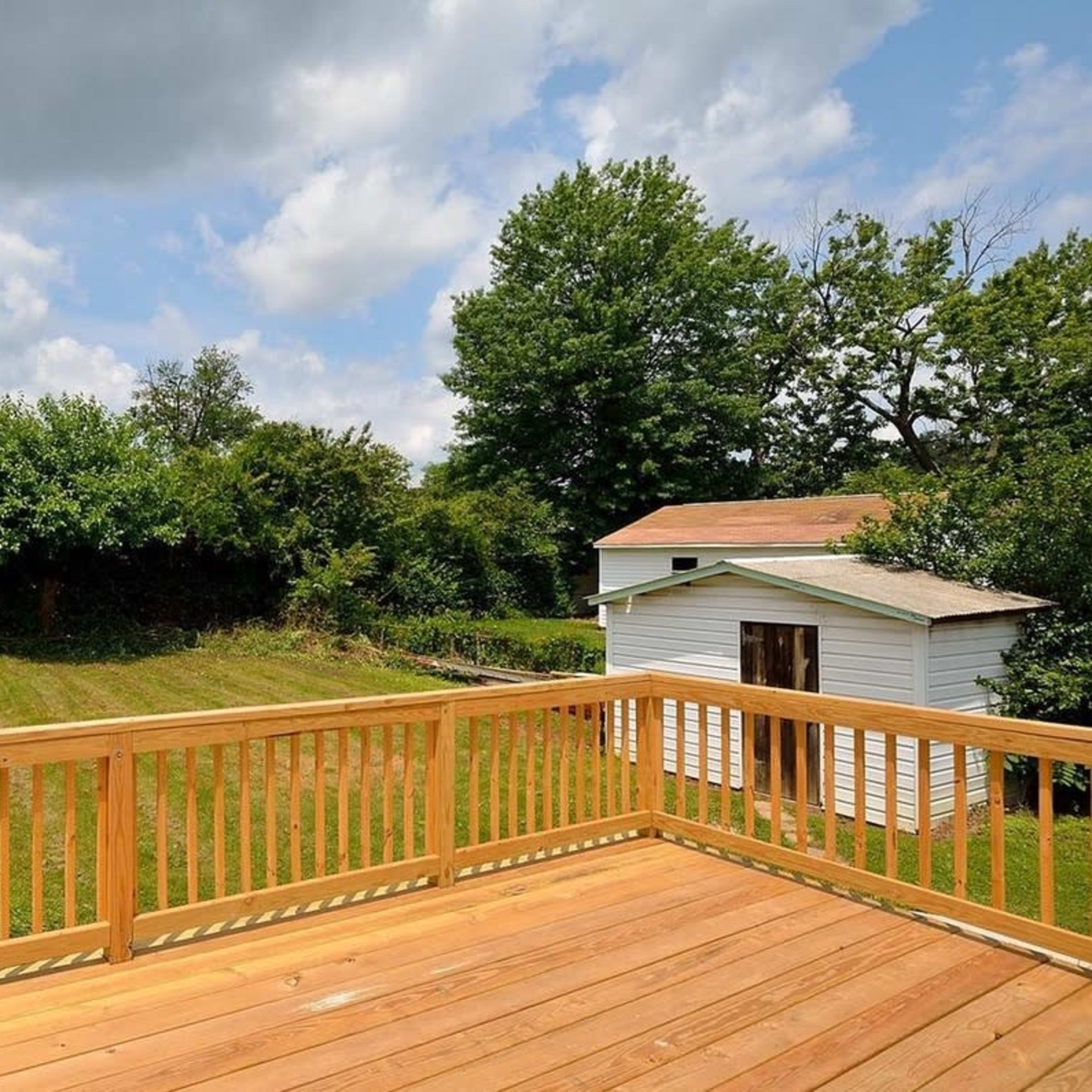 A wooden deck overlooks a grassy backyard with a small shed and trees in the background under a cloudy sky.