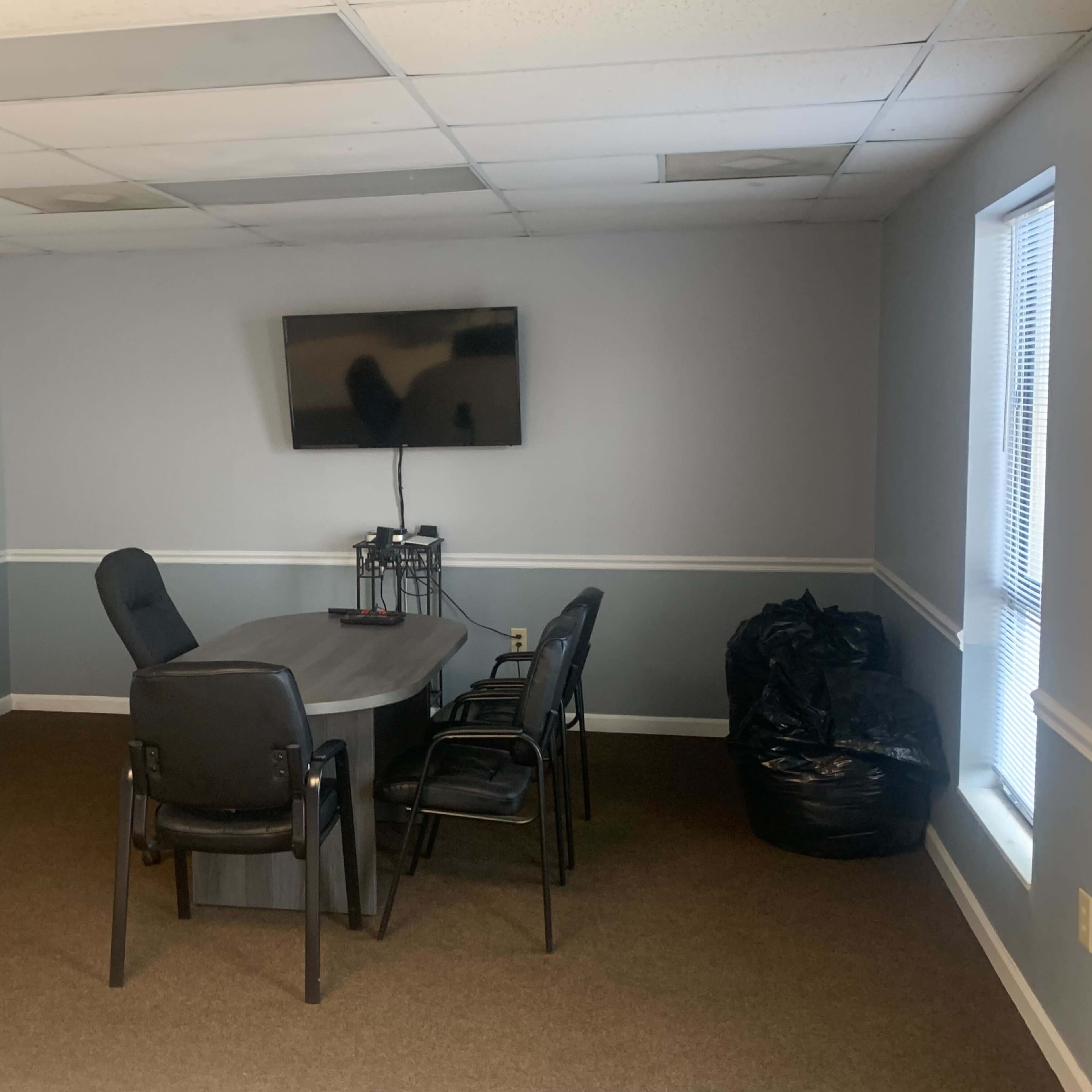 A sparsely furnished meeting room with a gray table, four black chairs, a television mounted on the wall, and a whiteboard.