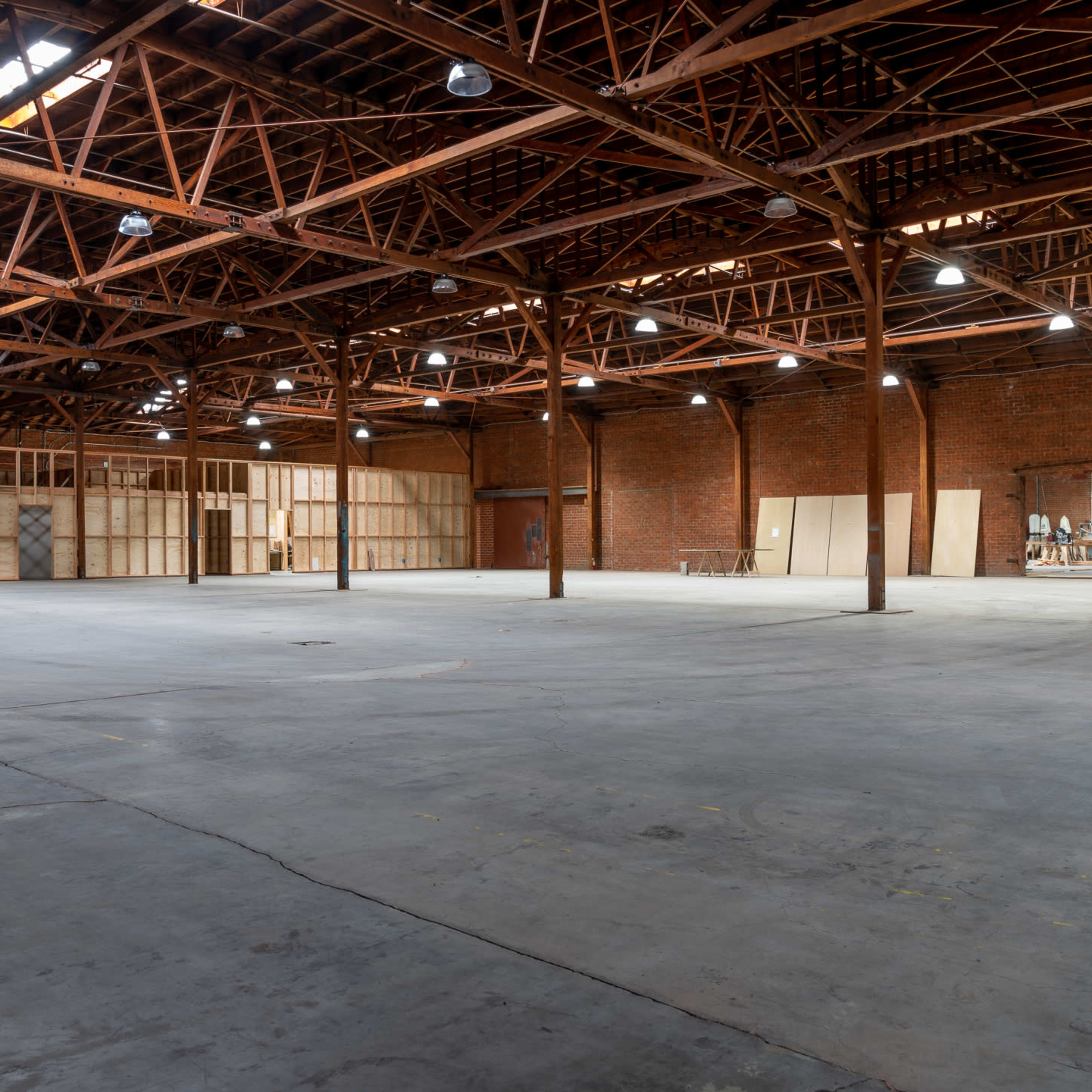 An empty warehouse with exposed brick walls and wooden beams, featuring concrete flooring and partitions made of wood in the background.
