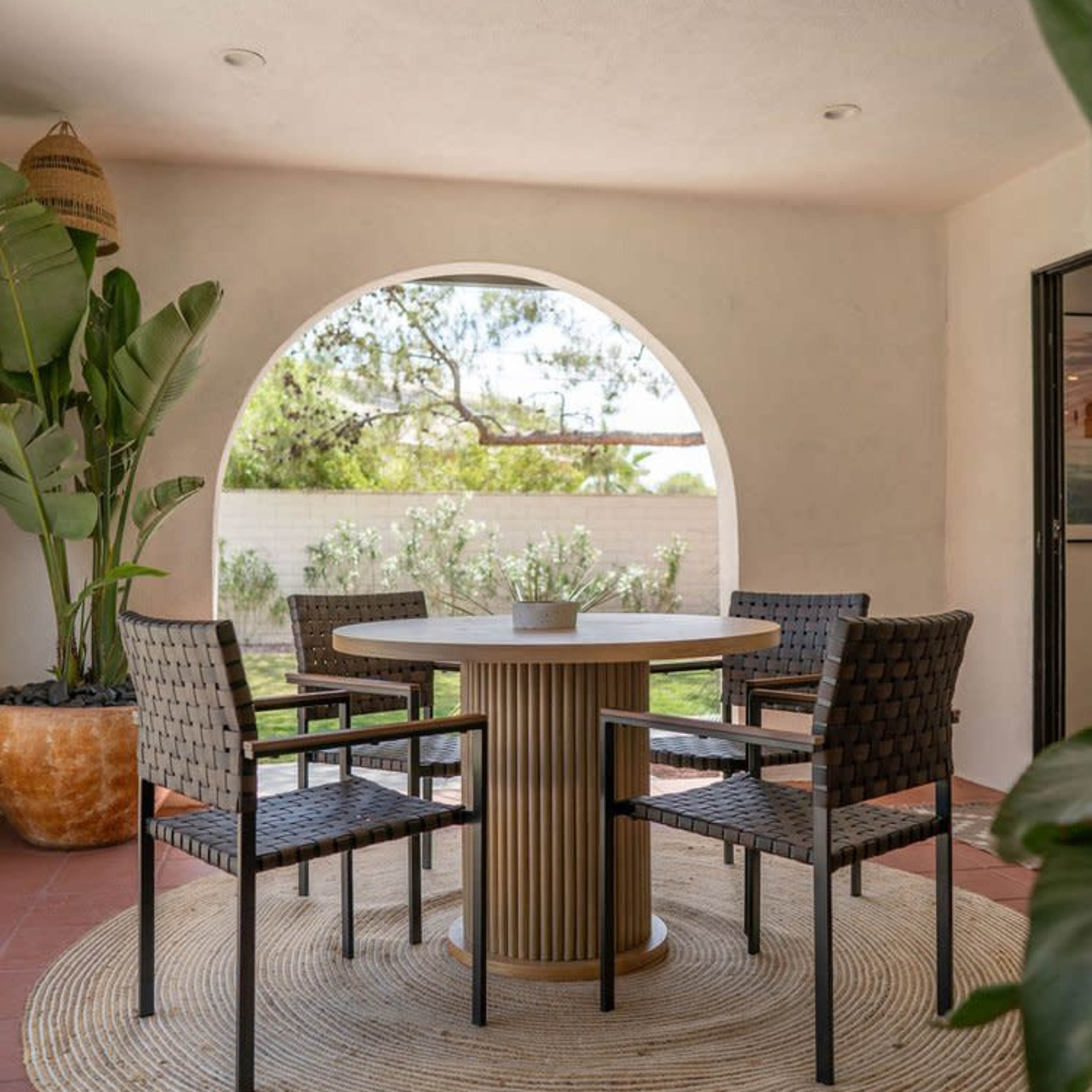 A round table with four chairs is positioned on a circular rug under an archway that leads to a garden view.