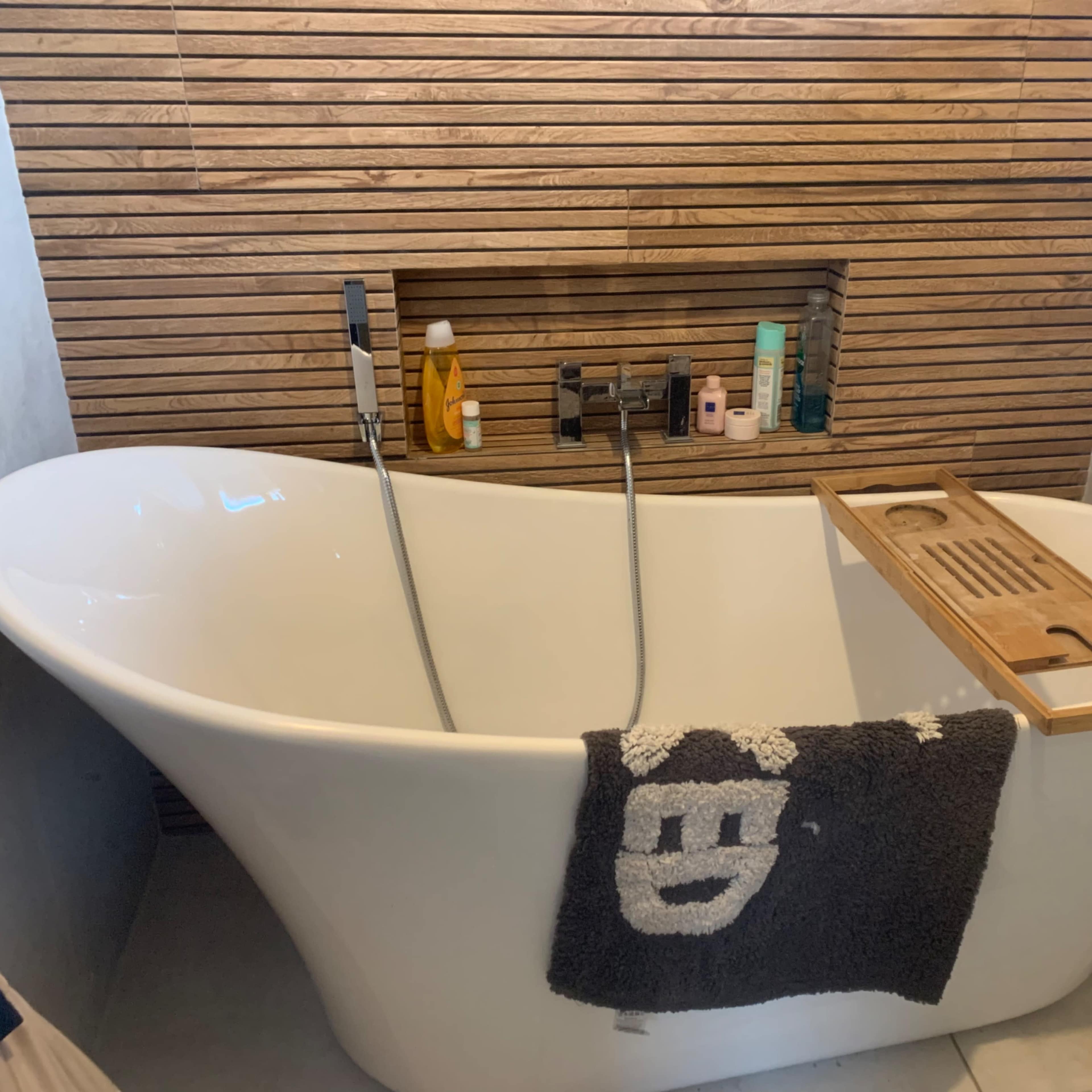 A modern bathtub is positioned against a wooden paneled wall, featuring a wooden bath tray and a black towel draped over the side.