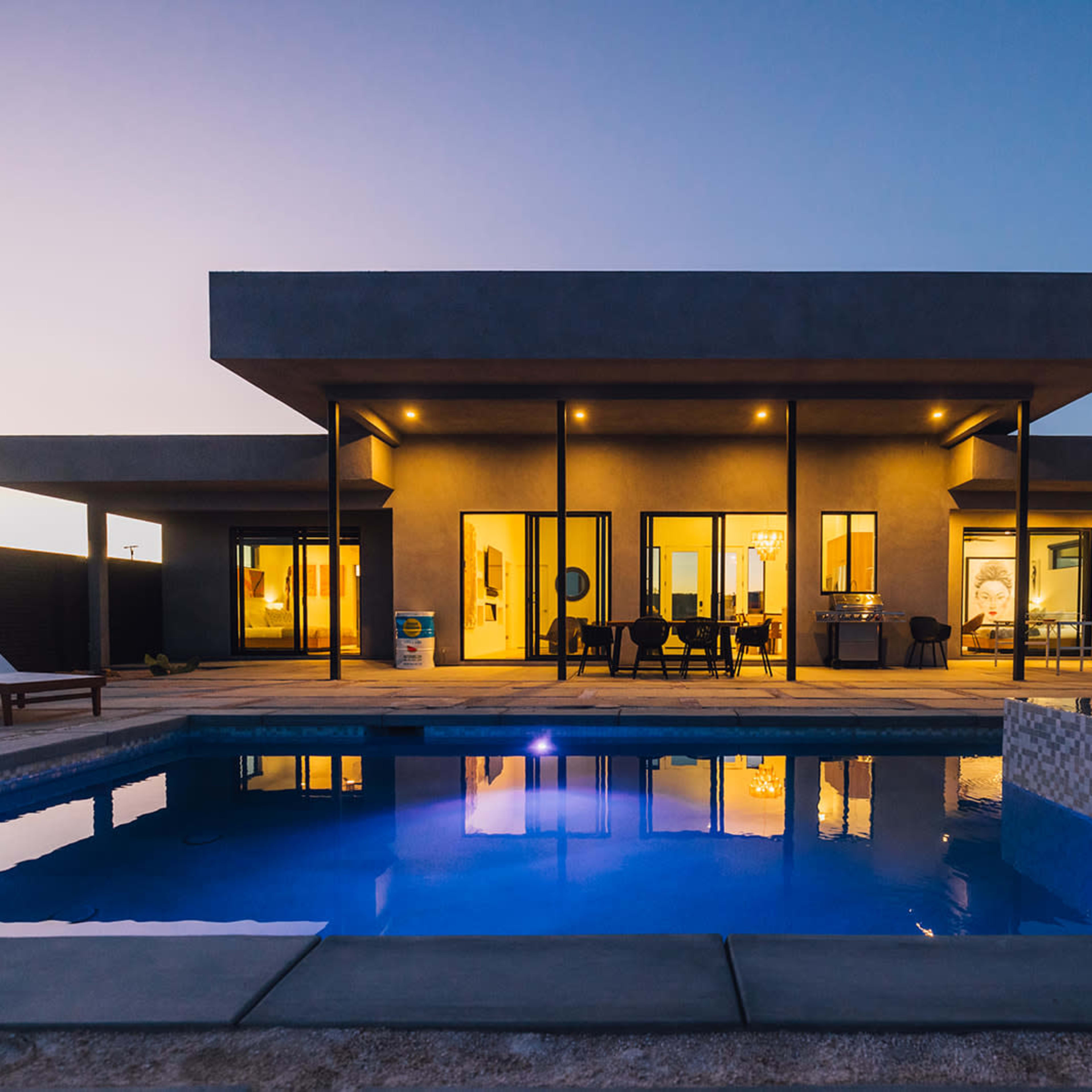 A modern house with large windows overlooks a swimming pool at dusk.