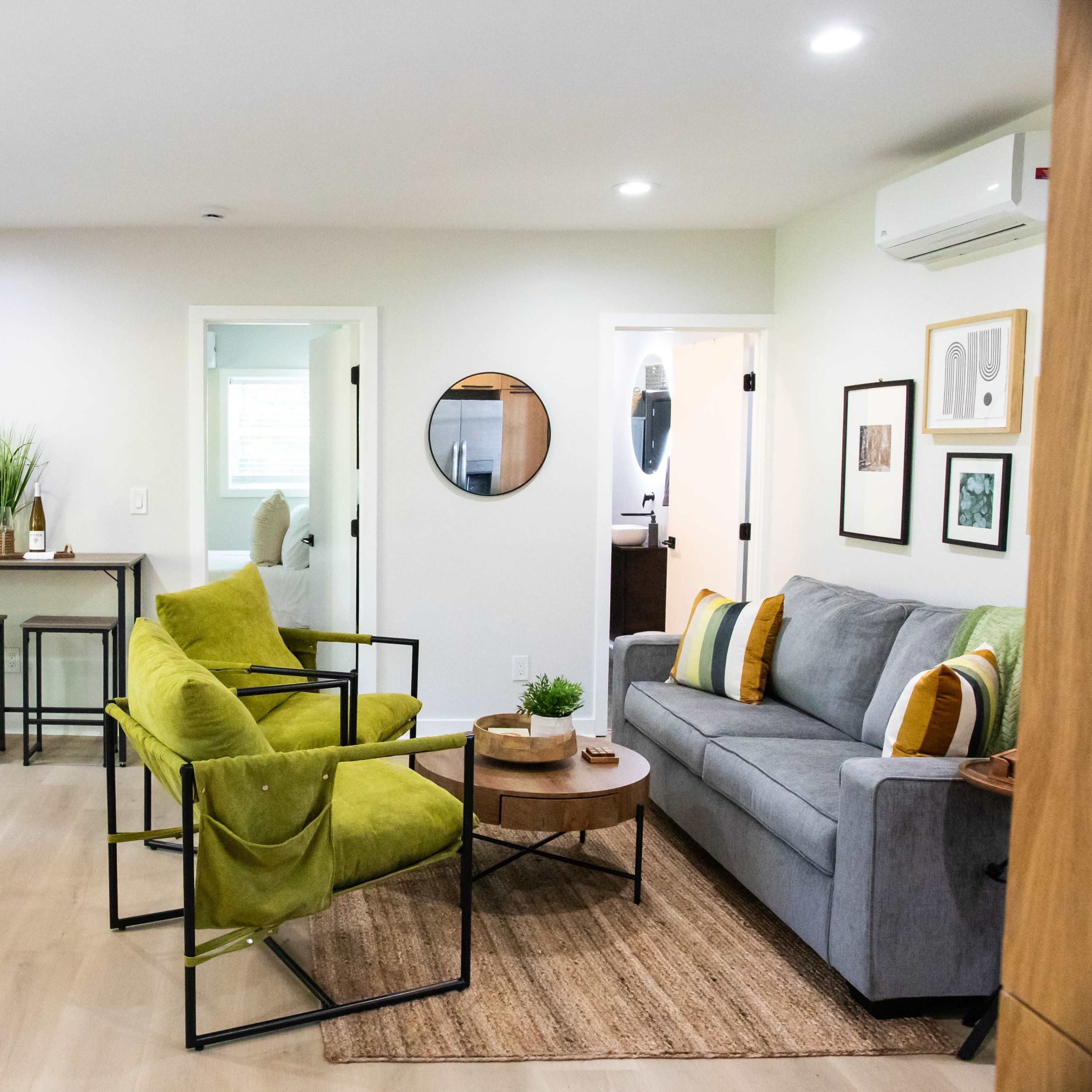 A modern living area featuring a gray sofa, two green chairs, a small dining table, and a flat-screen TV mounted on the wall.