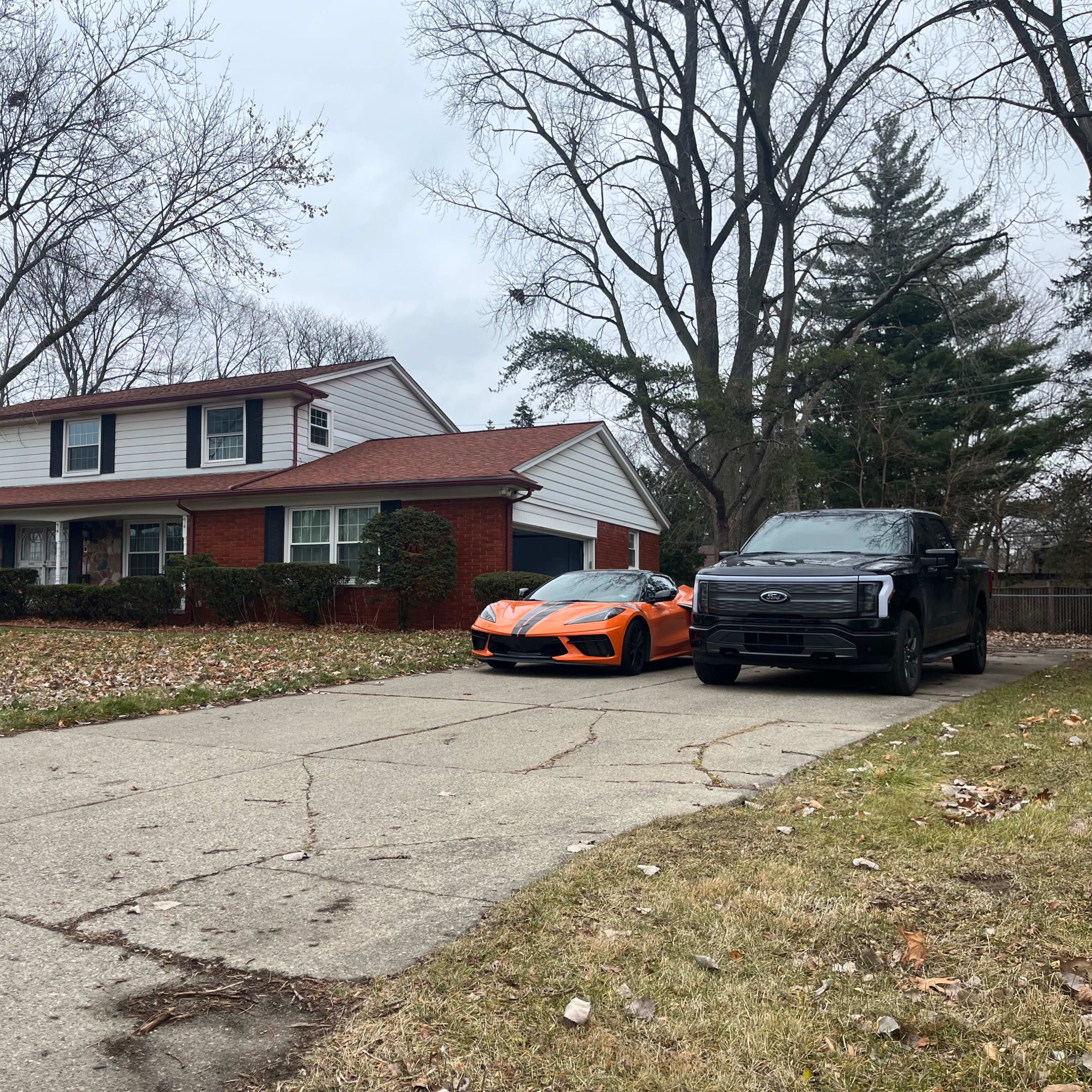 A two-story house with a red brick exterior sits next to an orange sports car and a black pickup truck on a driveway lined with uneven concrete.