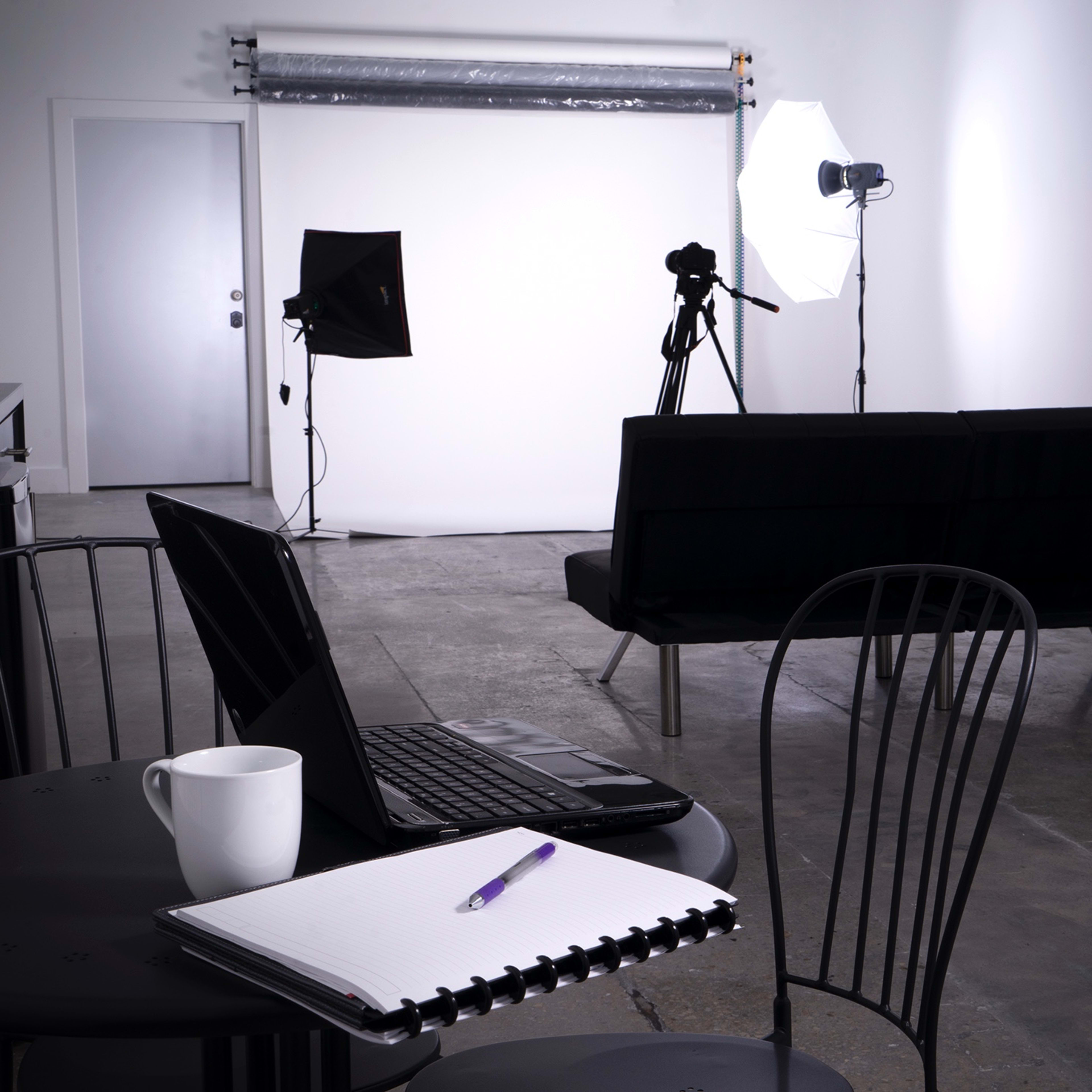 A photography studio is set up with a backdrop, lighting equipment, a camera on a tripod, a black sofa, and a table with a laptop and a notebook.