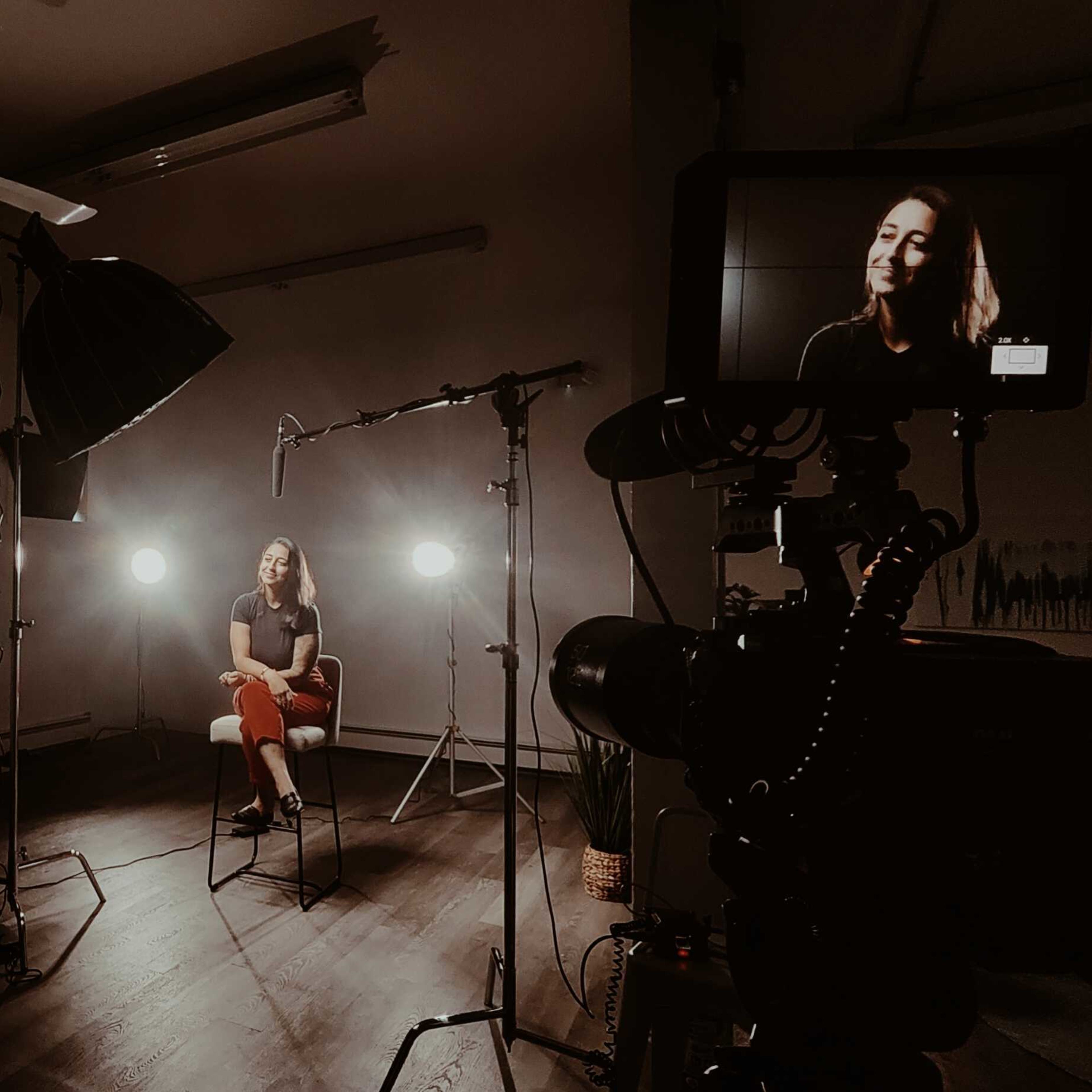 A woman sits on a chair in a well-lit studio, surrounded by video equipment and lighting.