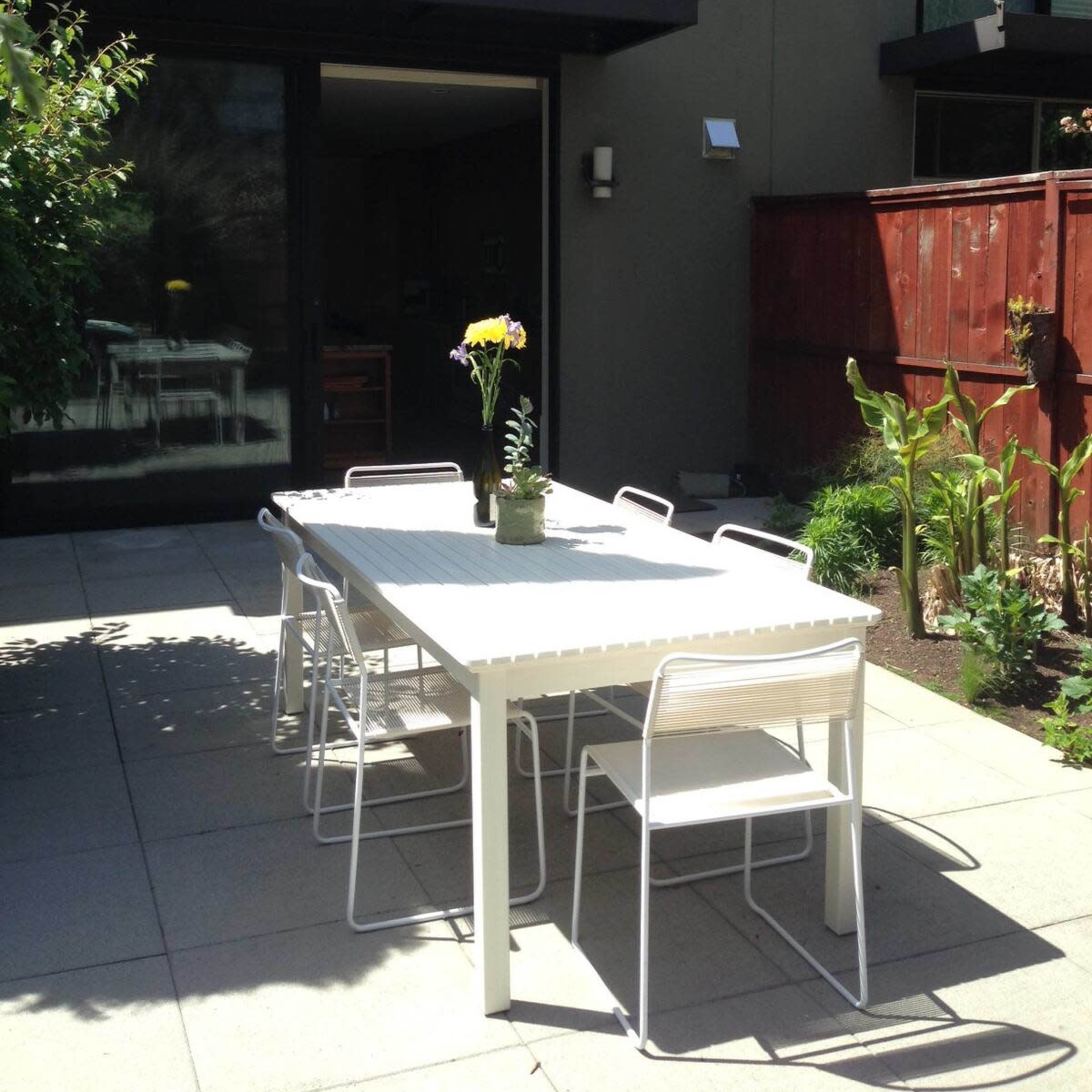 A white outdoor table with chairs sits in a patio area, surrounded by greenery and a wooden fence, in front of a modern building with large windows.
