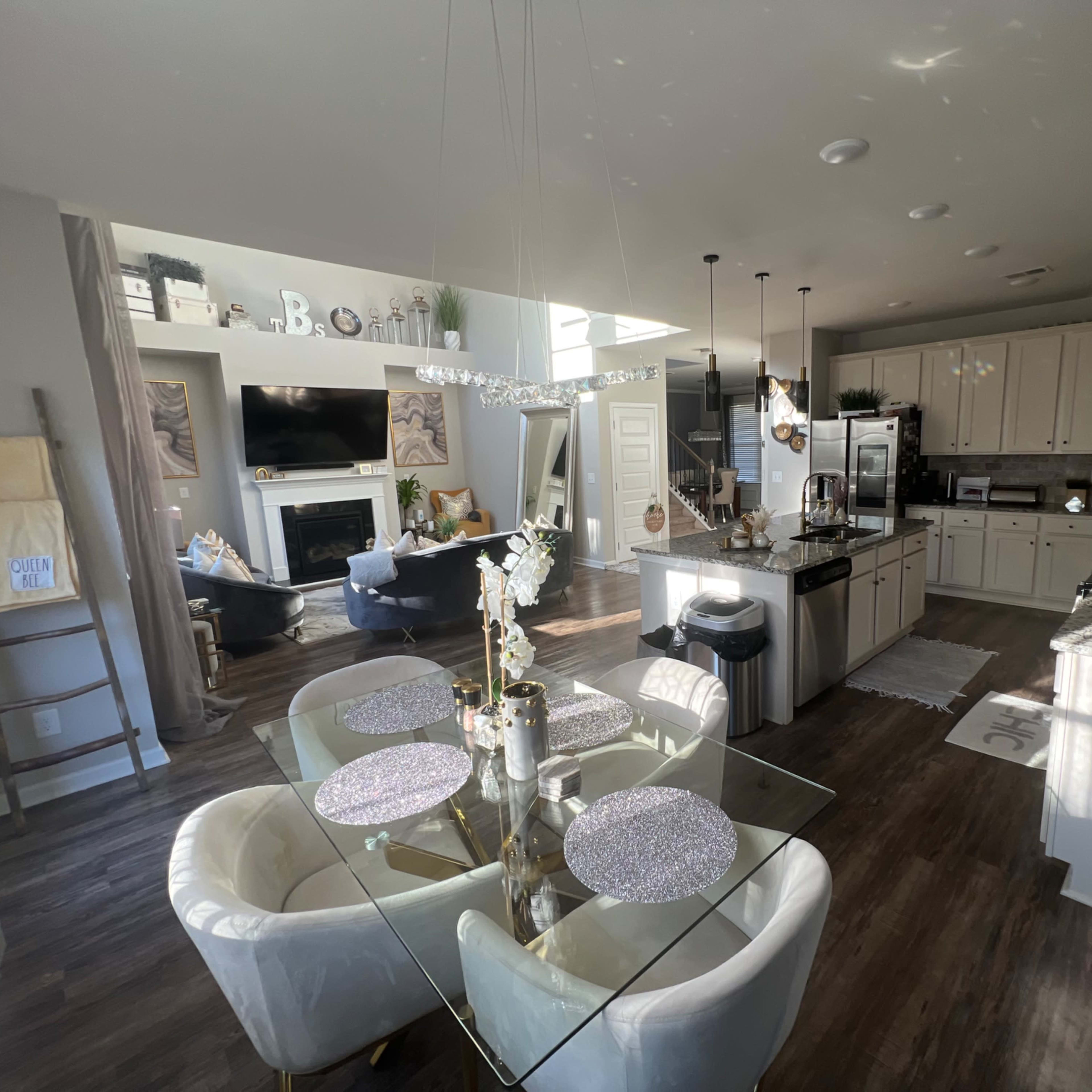 A modern kitchen and dining area with a glass table, light-colored cabinets, and a living room visible in the background.