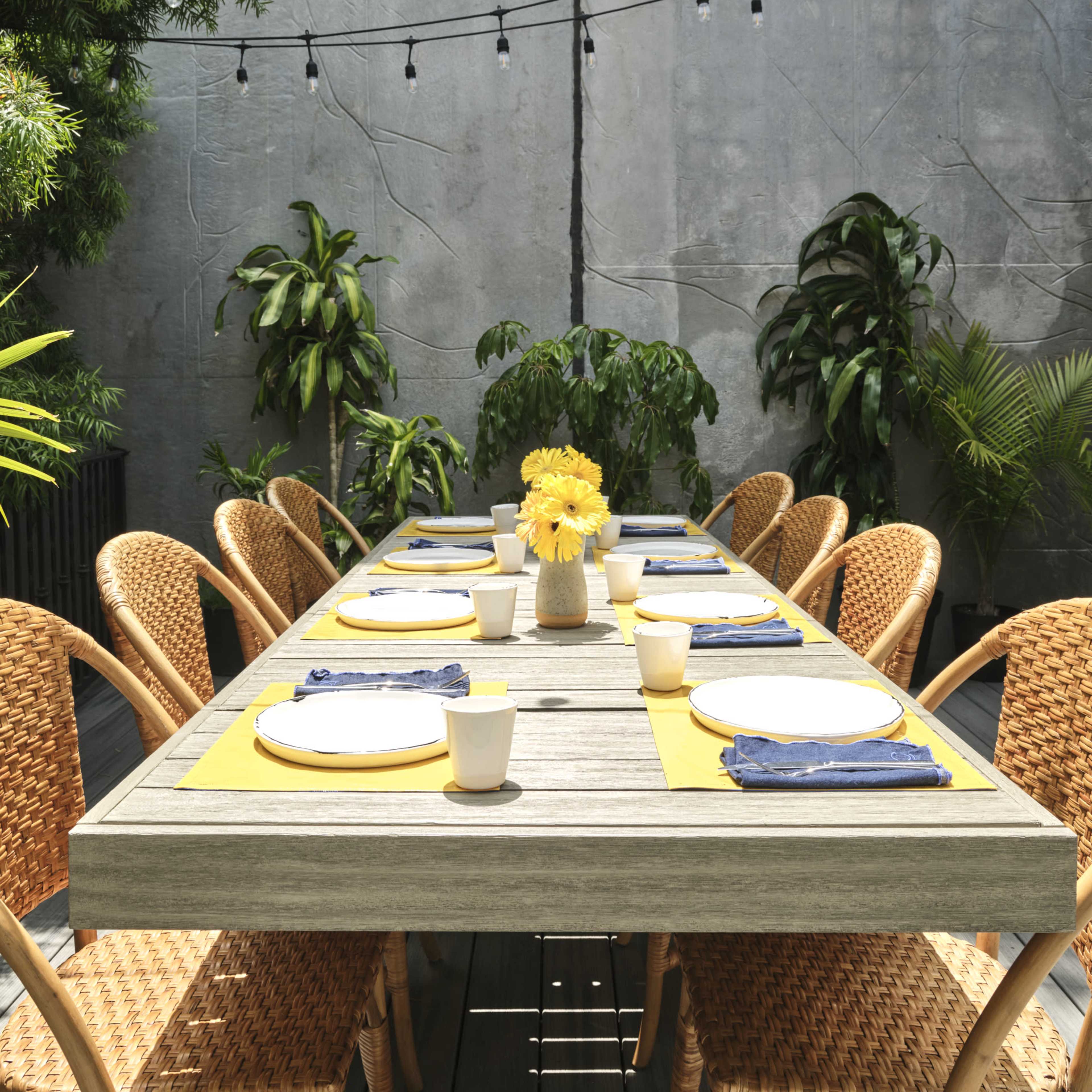 A long wooden dining table is set with plates and cups, surrounded by woven chairs in a garden area adorned with plants and string lights overhead.