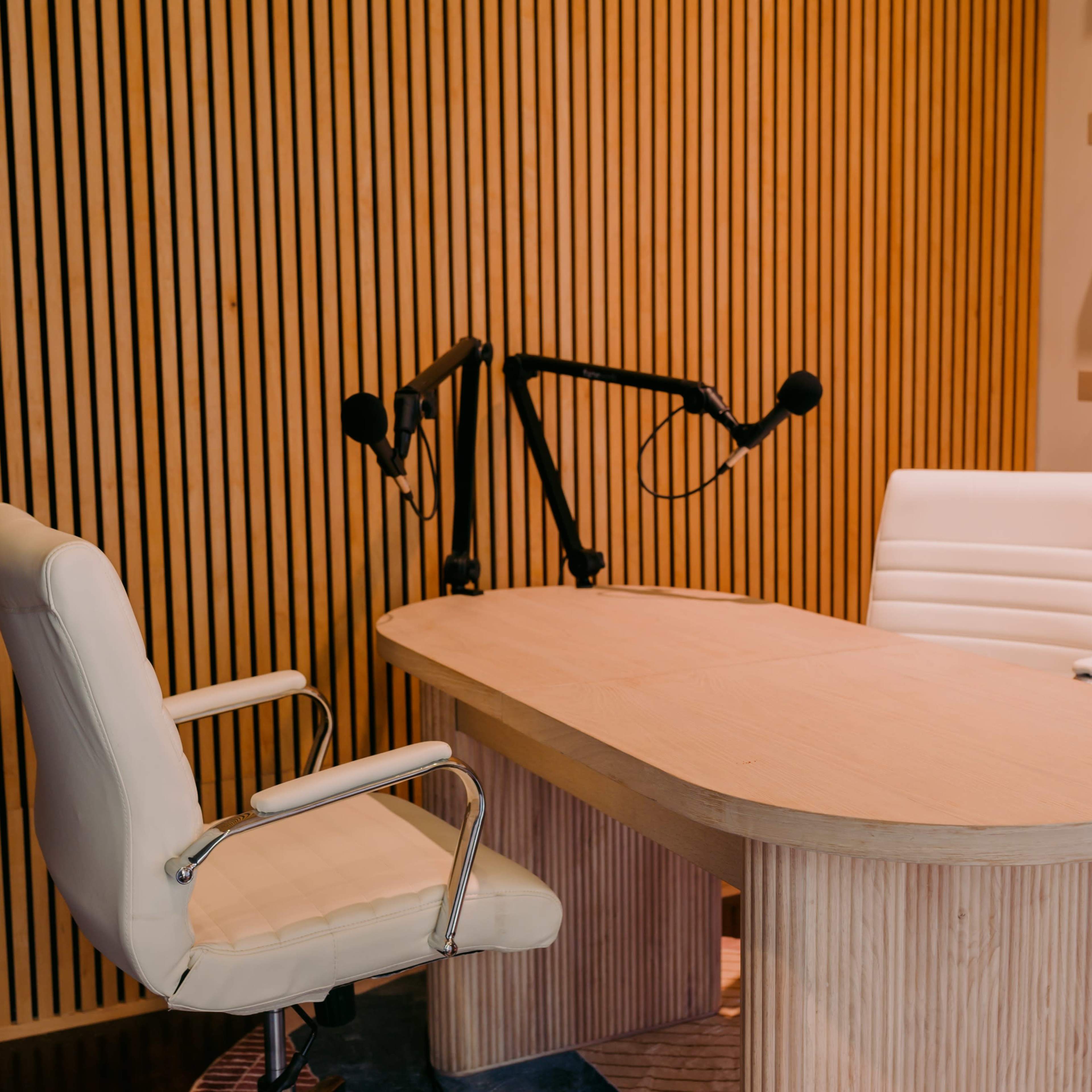 A minimalistic podcast studio featuring a wooden table, two white chairs, and a wooden wall with vertical slats.