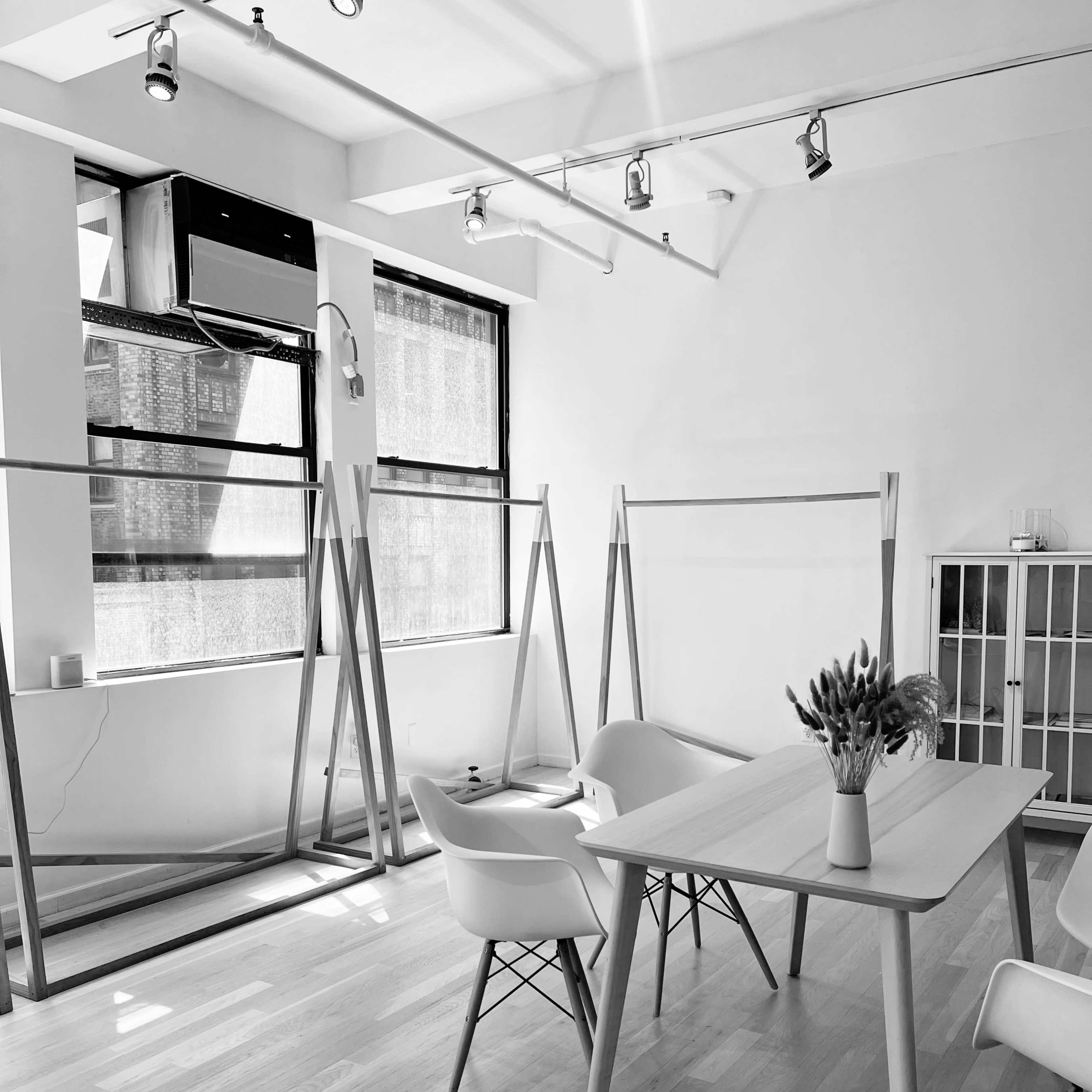 A minimalist interior space with two large windows, several clothing racks, a table with a vase of flowers, and a small cabinet.