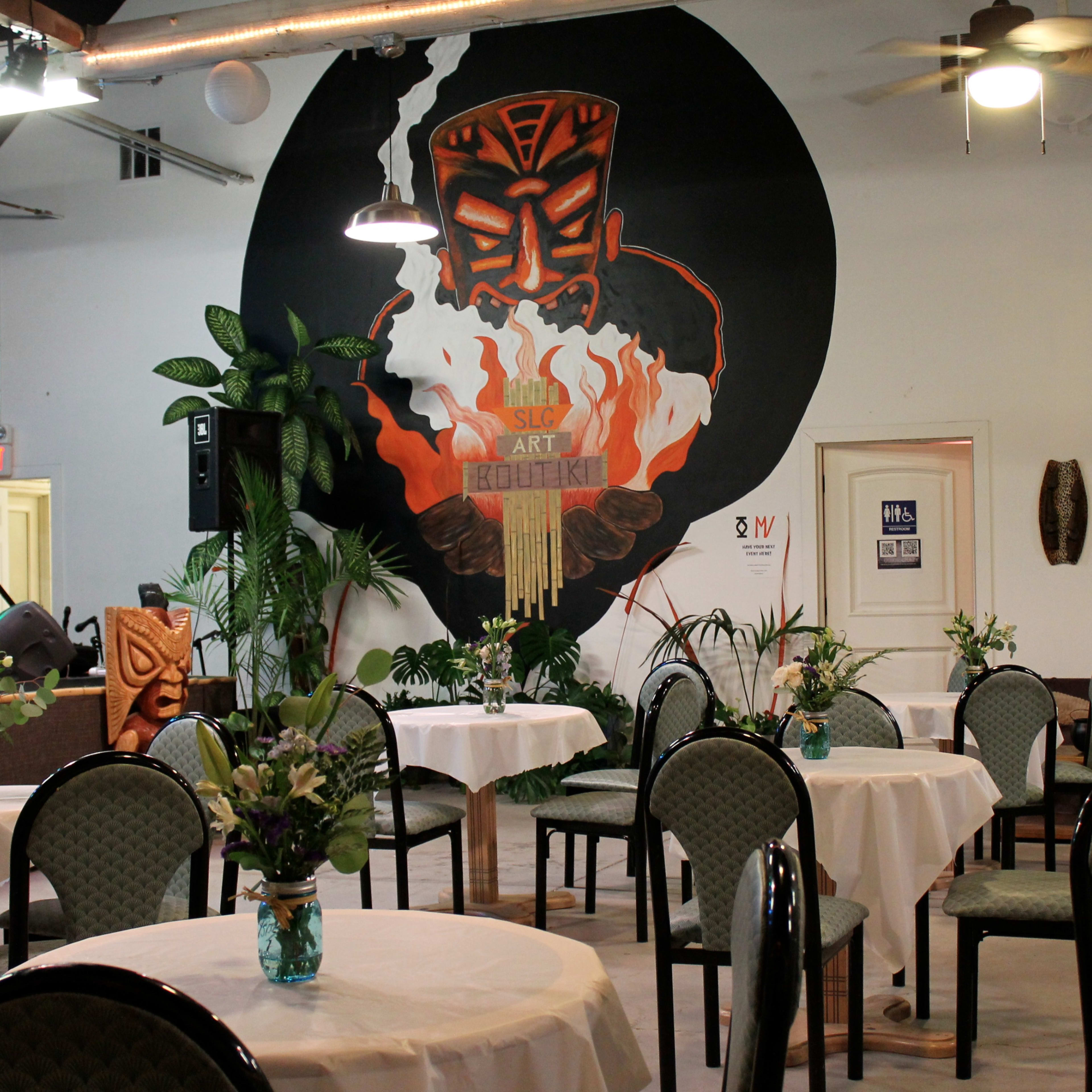 A decorated event space with tables set for dining, featuring a large mural of a Tiki figure on the wall.