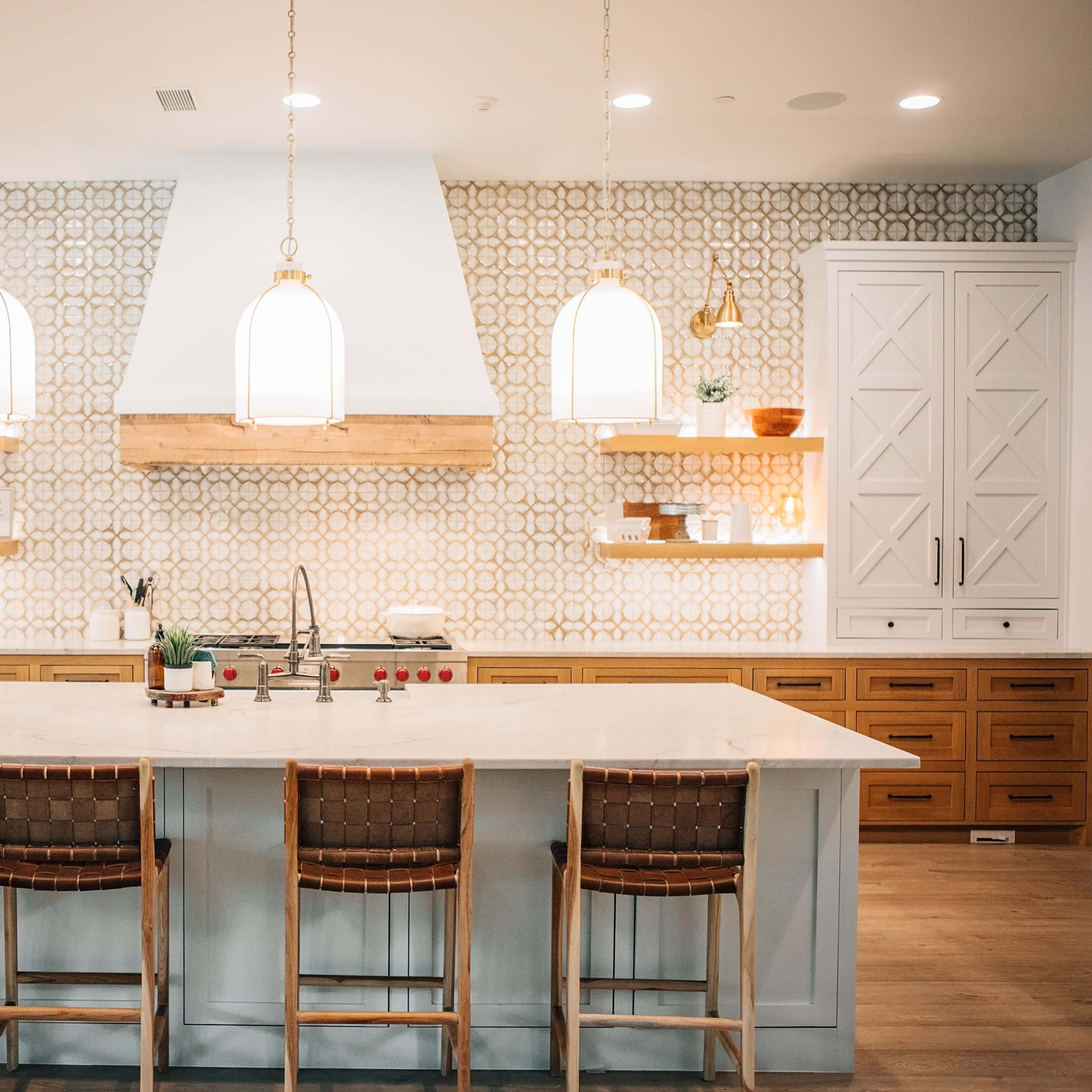 A modern kitchen features a large white island with wooden bar stools, decorative wall shelving, and patterned tile behind the stove.
