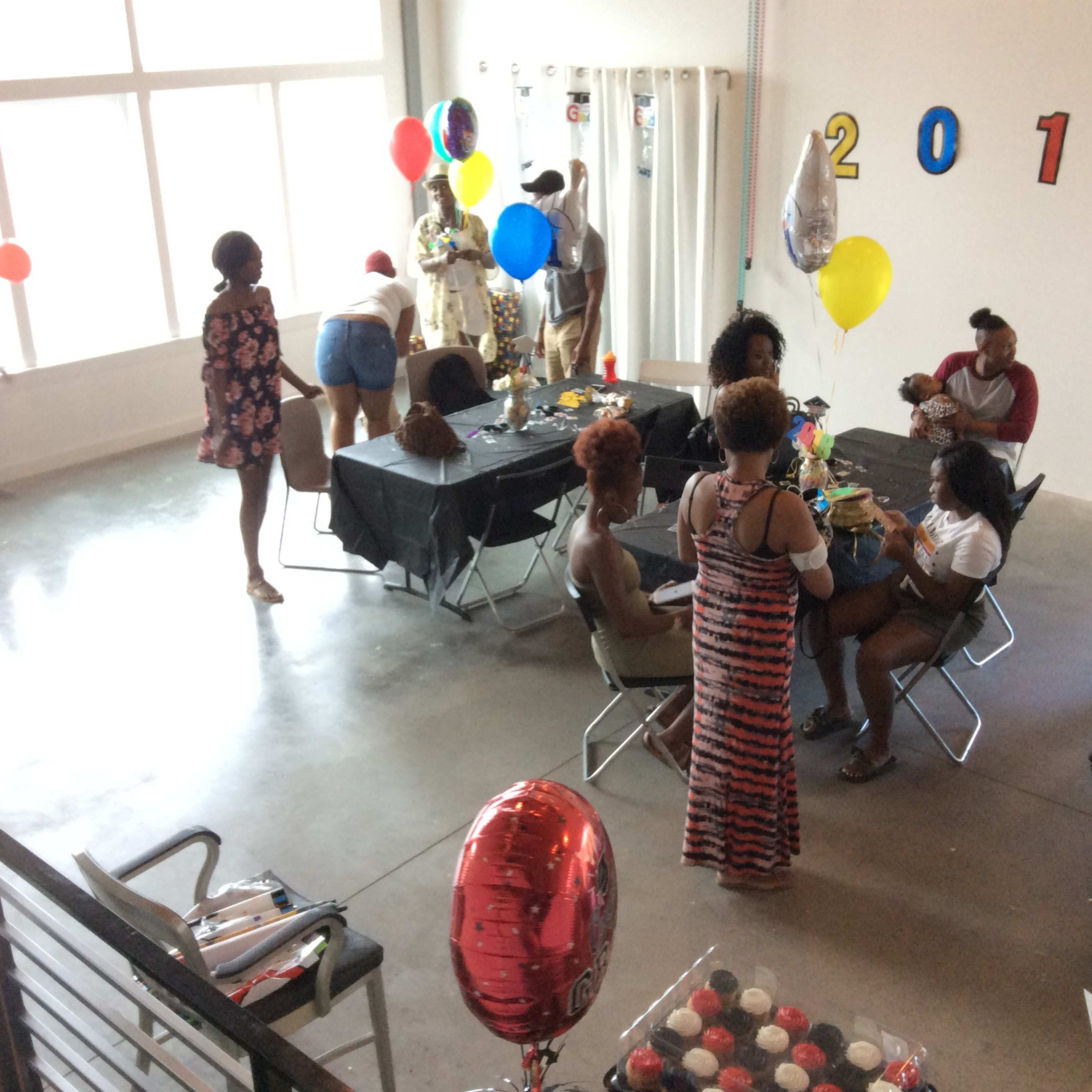 A group of people gather in a brightly lit room decorated with balloons and "2018" signage, engaged in a festive gathering around tables with food and drinks.