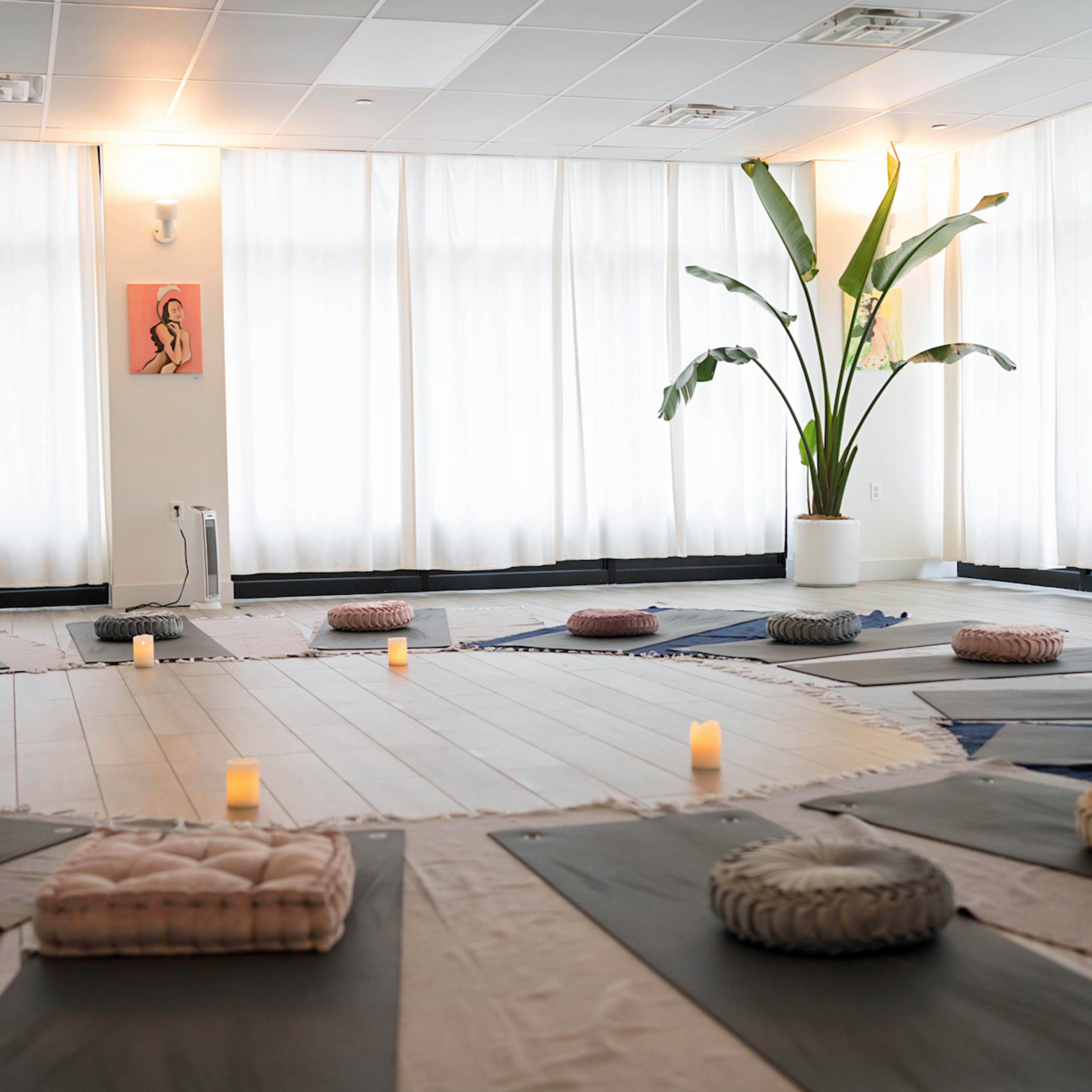 A tranquil yoga studio with meditation cushions arranged in a circular pattern around flickering candles and large windows with sheer curtains.