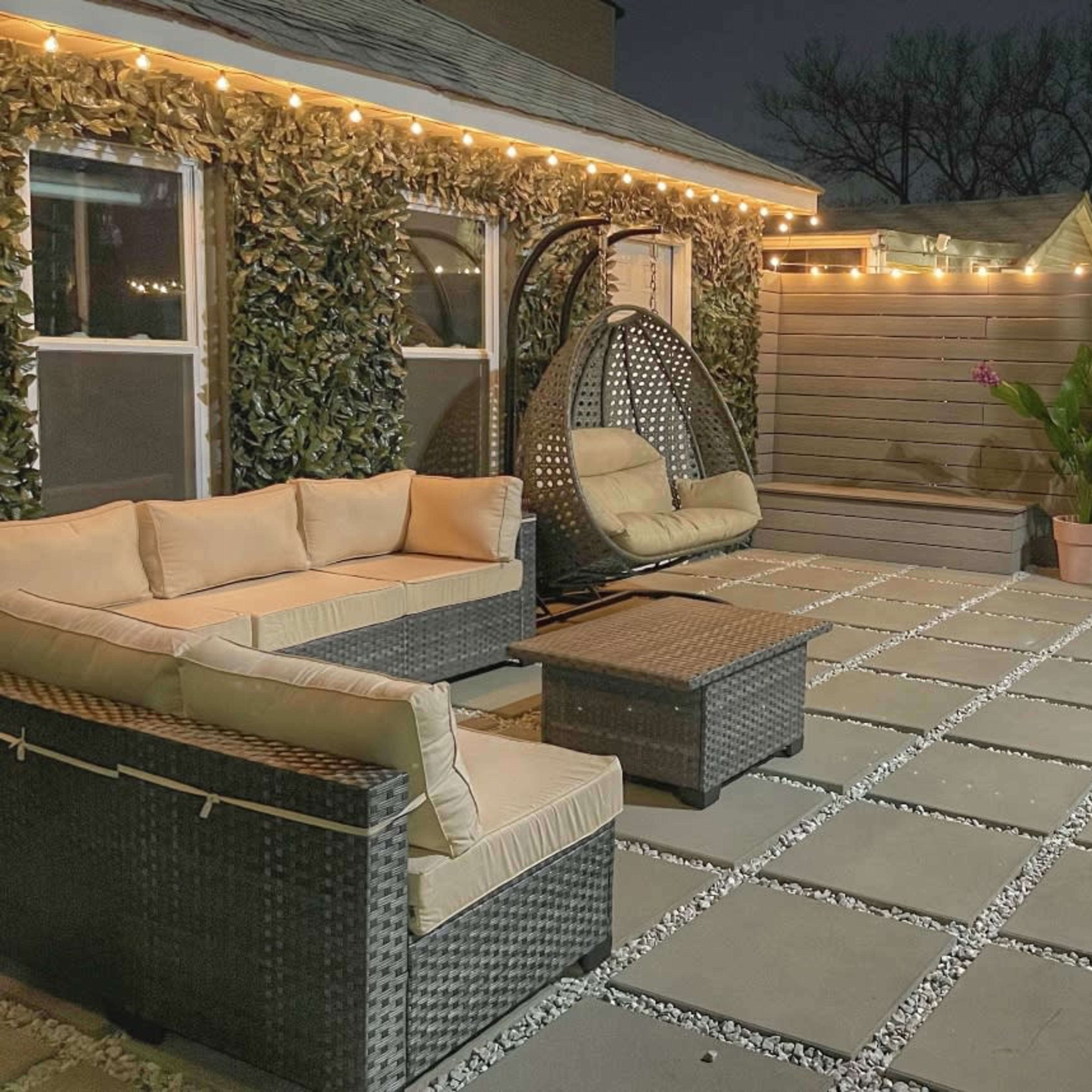 A cozy patio features a woven seating area with cushions, a hanging chair, and decorative lights illuminating the space at night.