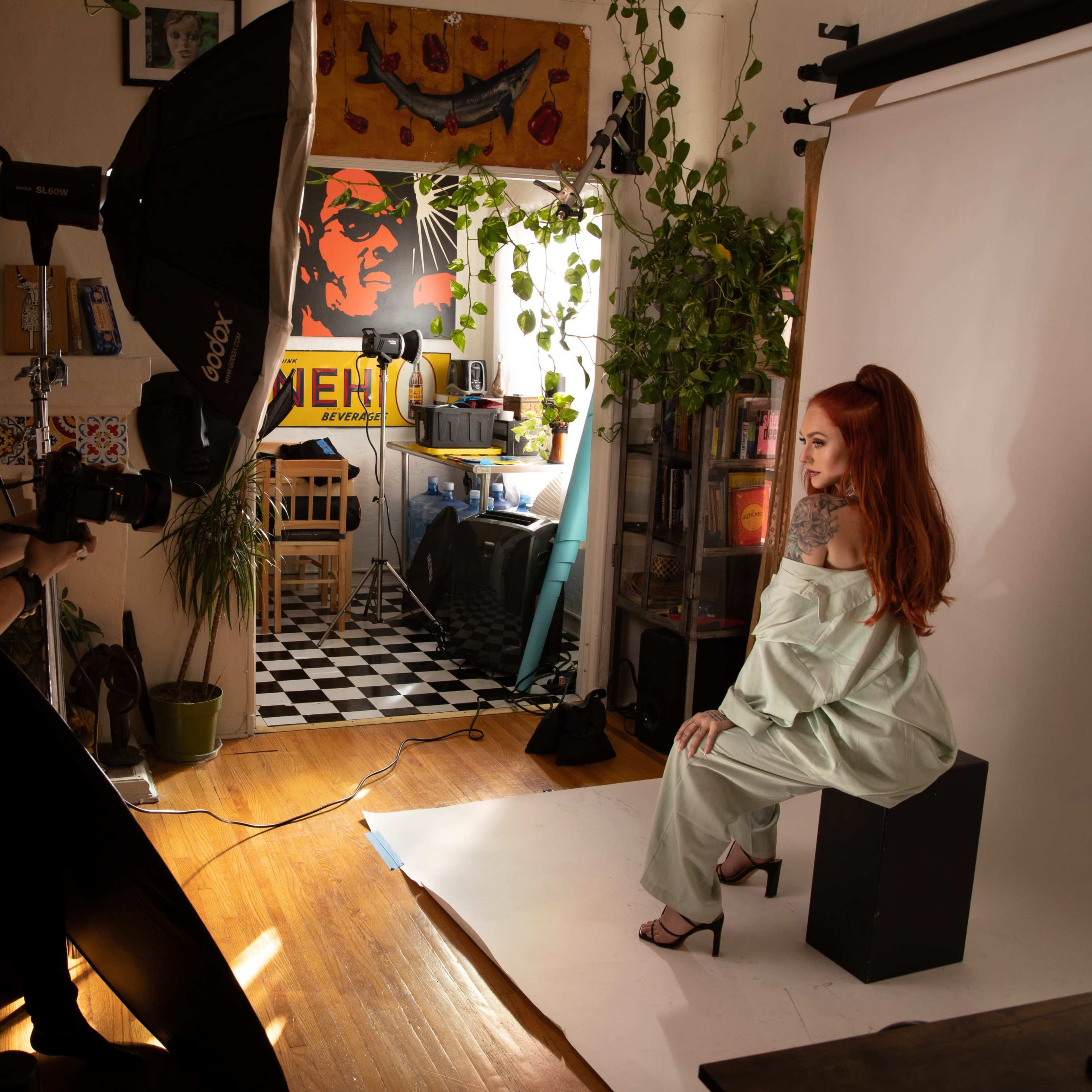 A photographer is taking a portrait of a woman seated on a stool in a well-decorated studio with a backdrop and natural light.