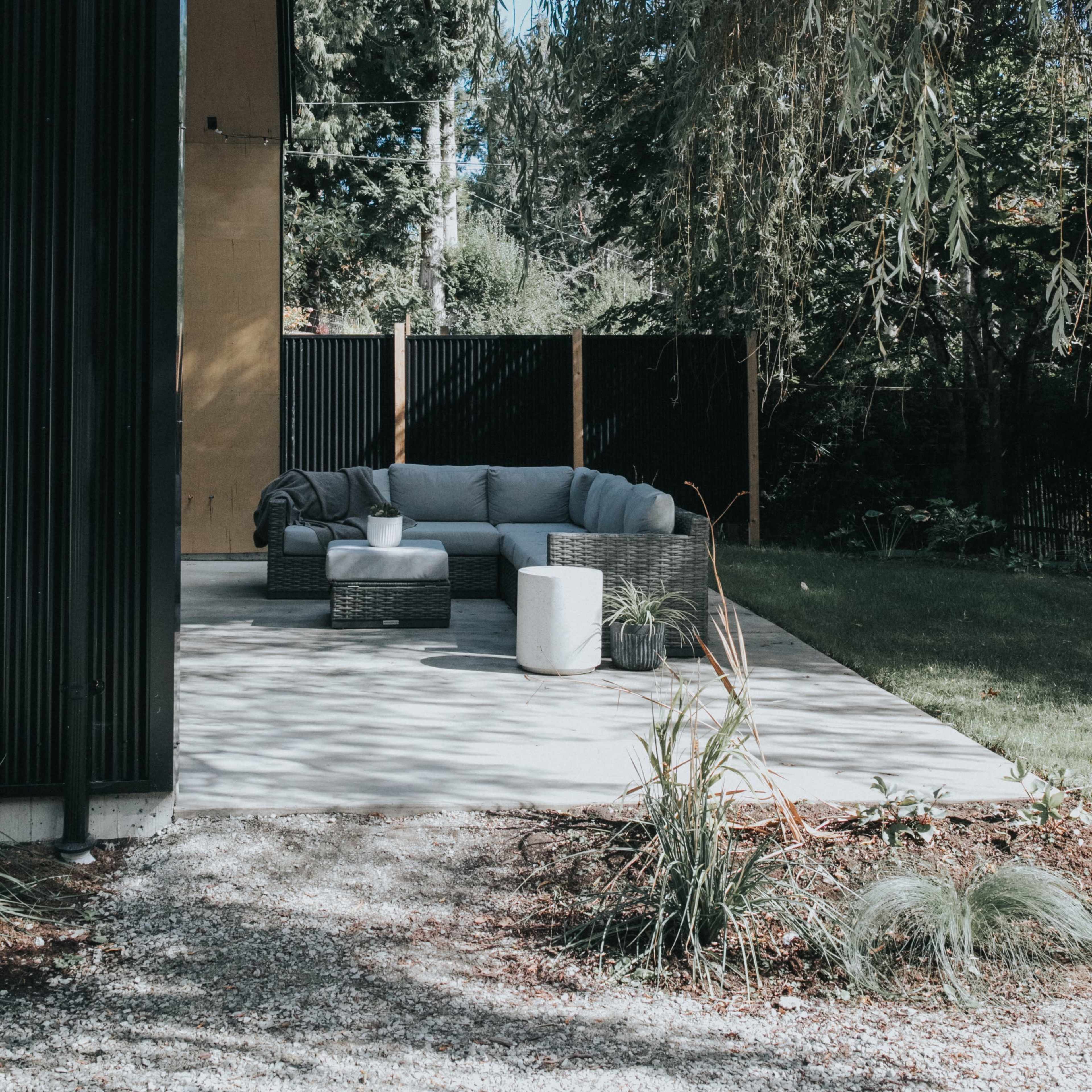 A modern patio with outdoor furniture is situated beside a house, bordered by trees and a black fence.