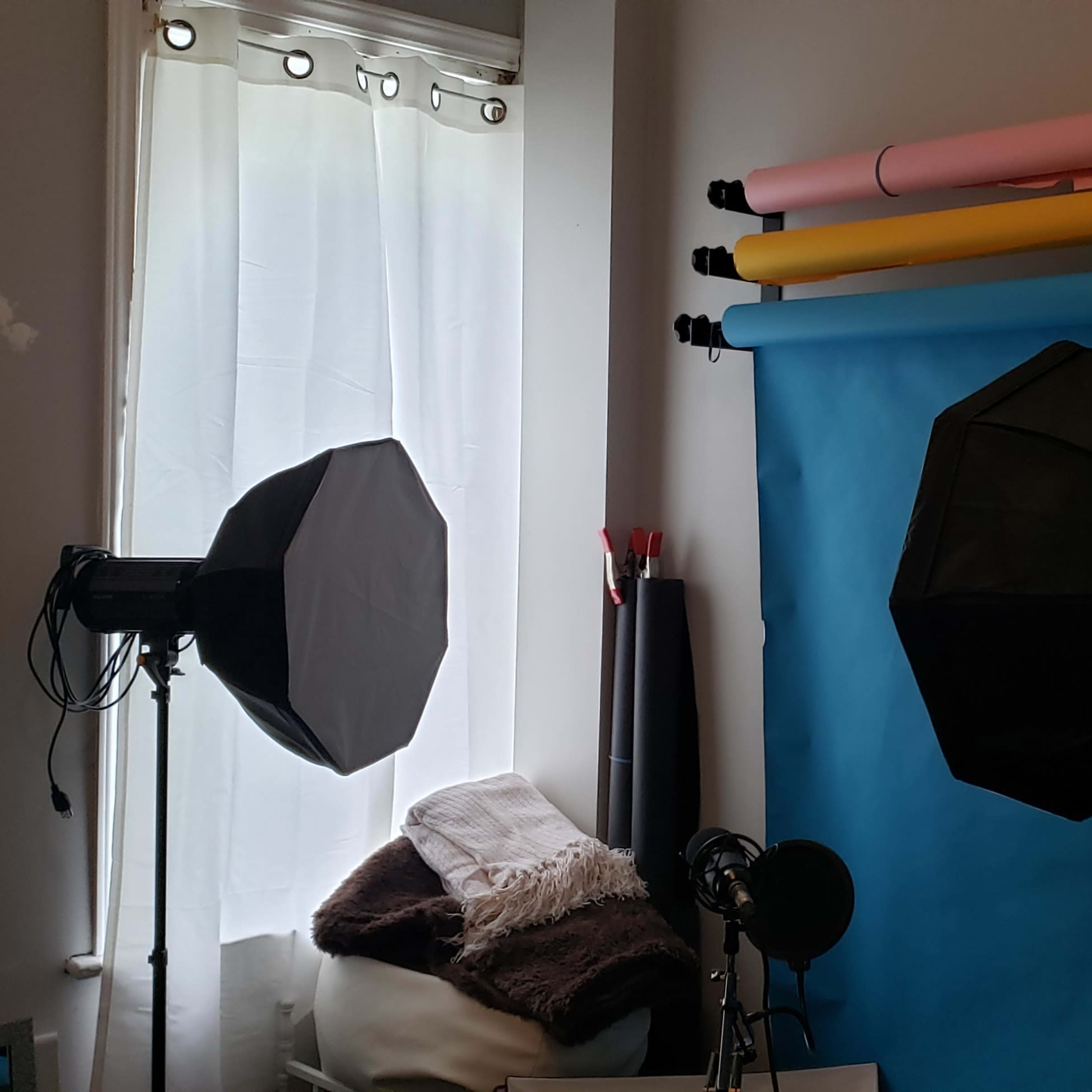 A photography setup with two softbox lights and a backdrop stand featuring rolls of colored paper against a wall, alongside a window with sheer curtains.