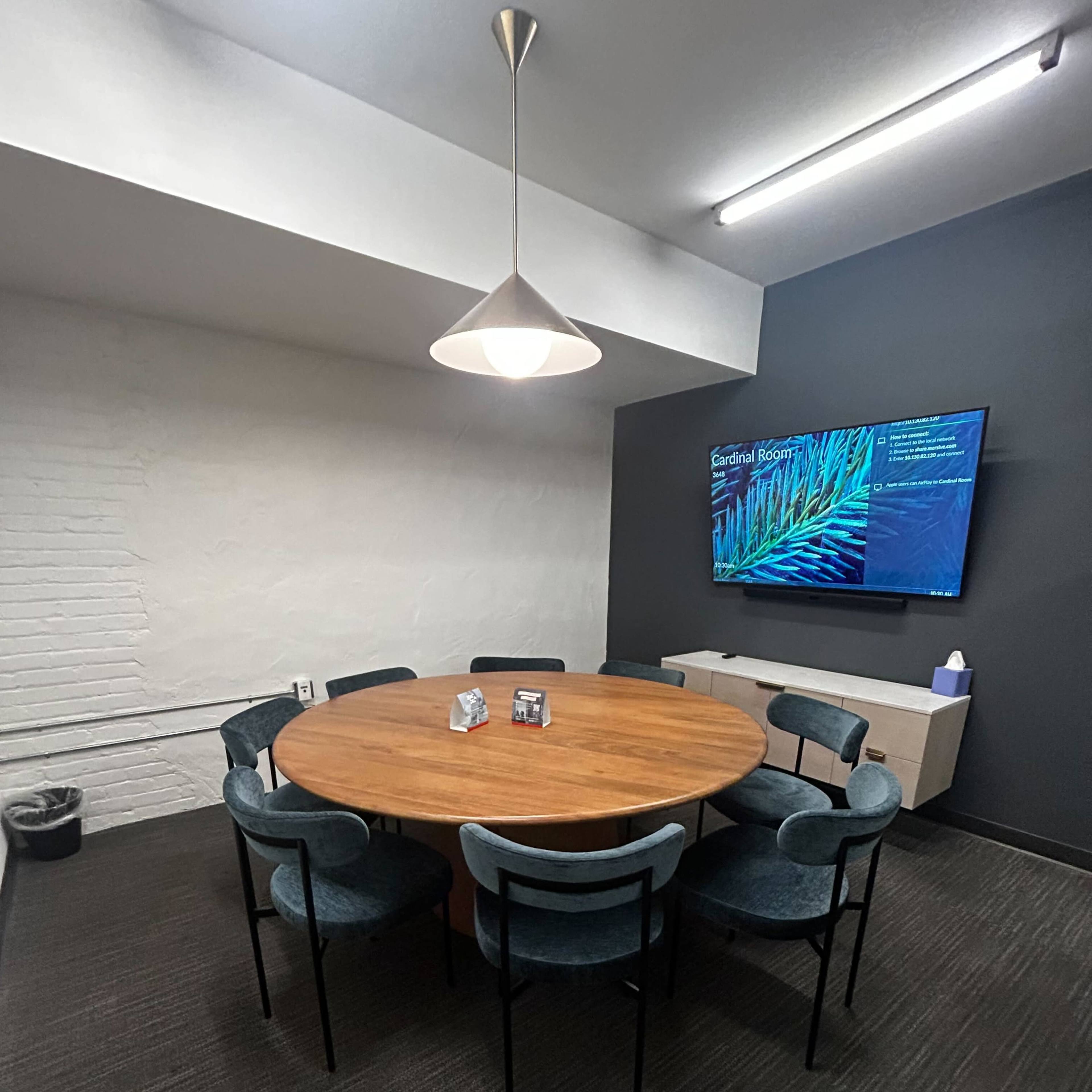 A circular wooden table surrounded by six upholstered chairs is set in a meeting room with a wall-mounted screen displaying information.