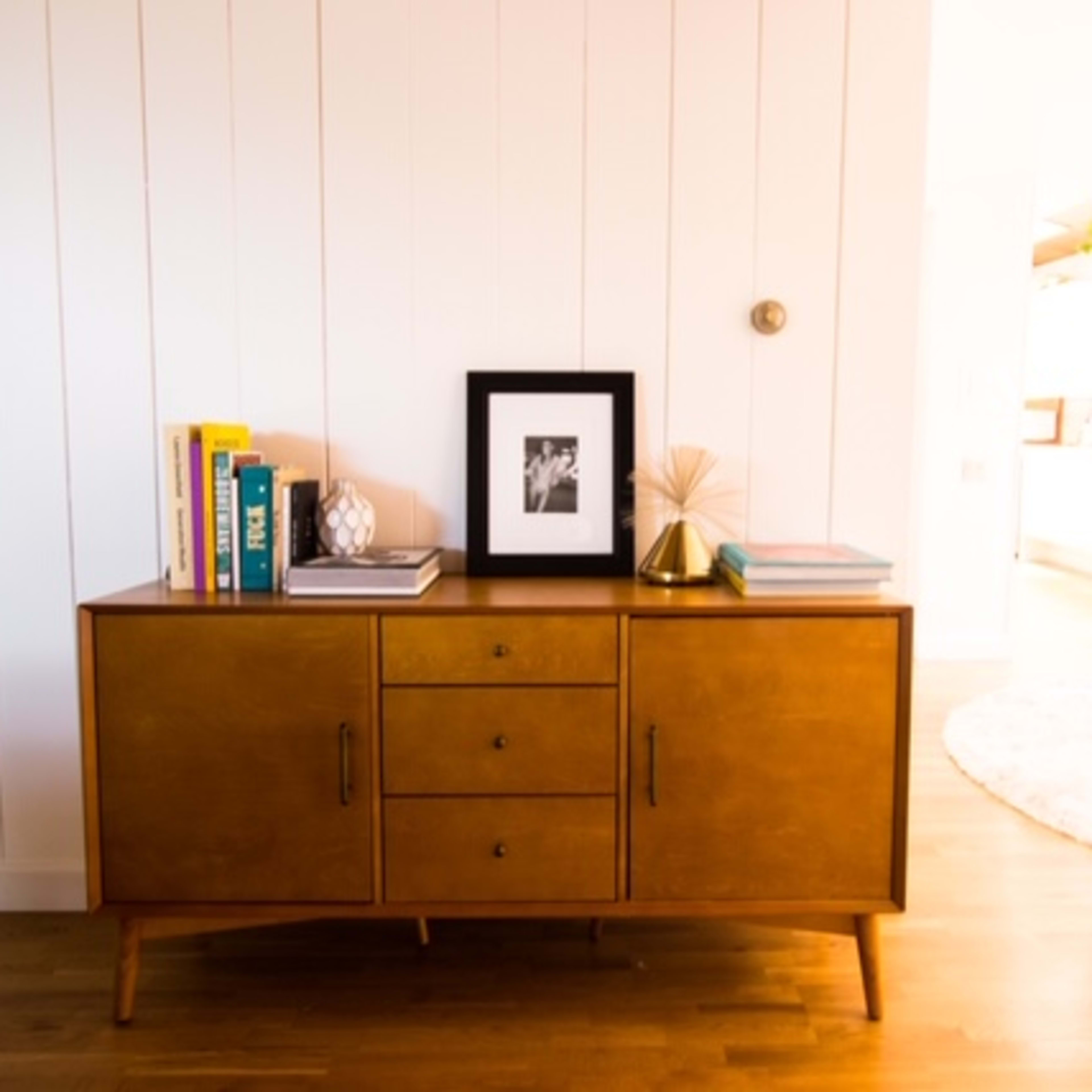 A mid-century modern wooden sideboard with three drawers and decorative items stands against a white-paneled wall, with a dining area visible in the background.