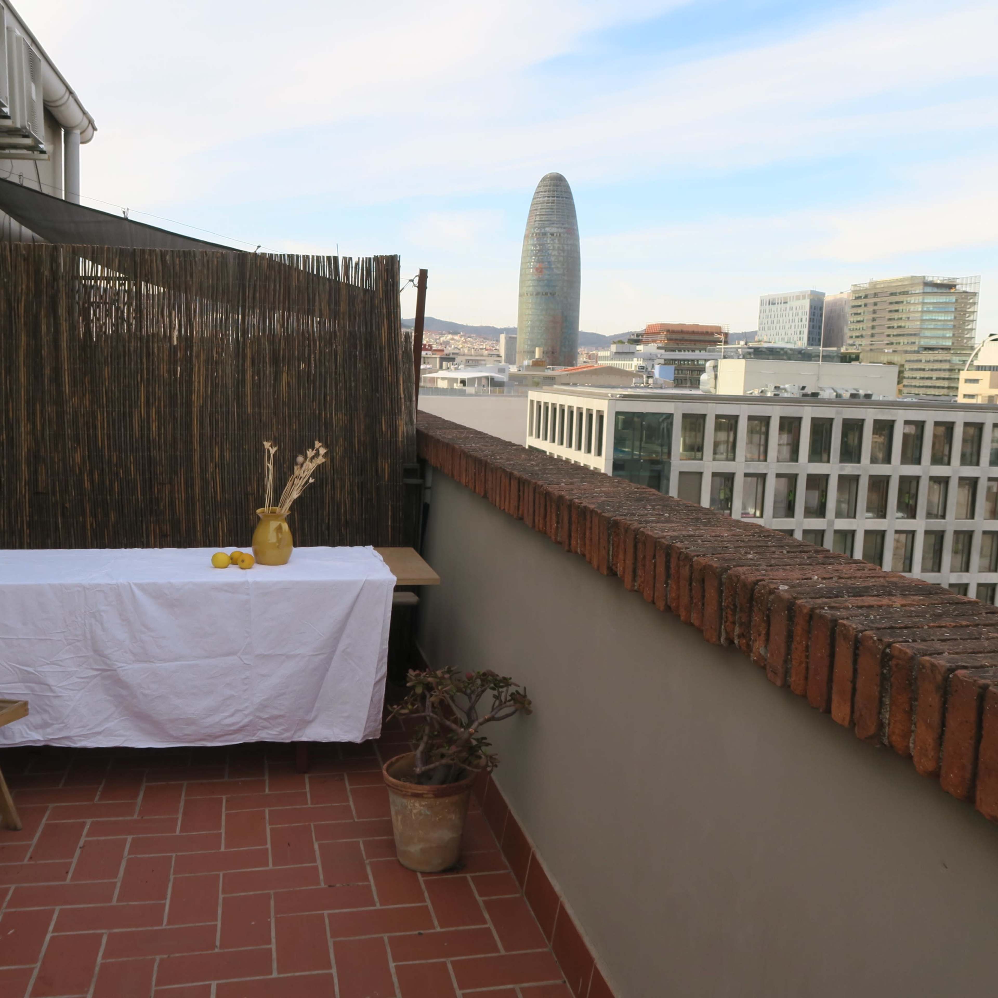 A balcony features a wooden table covered with a white cloth, a chair, and a backdrop of high-rise buildings, including a prominent skyscraper in the distance.