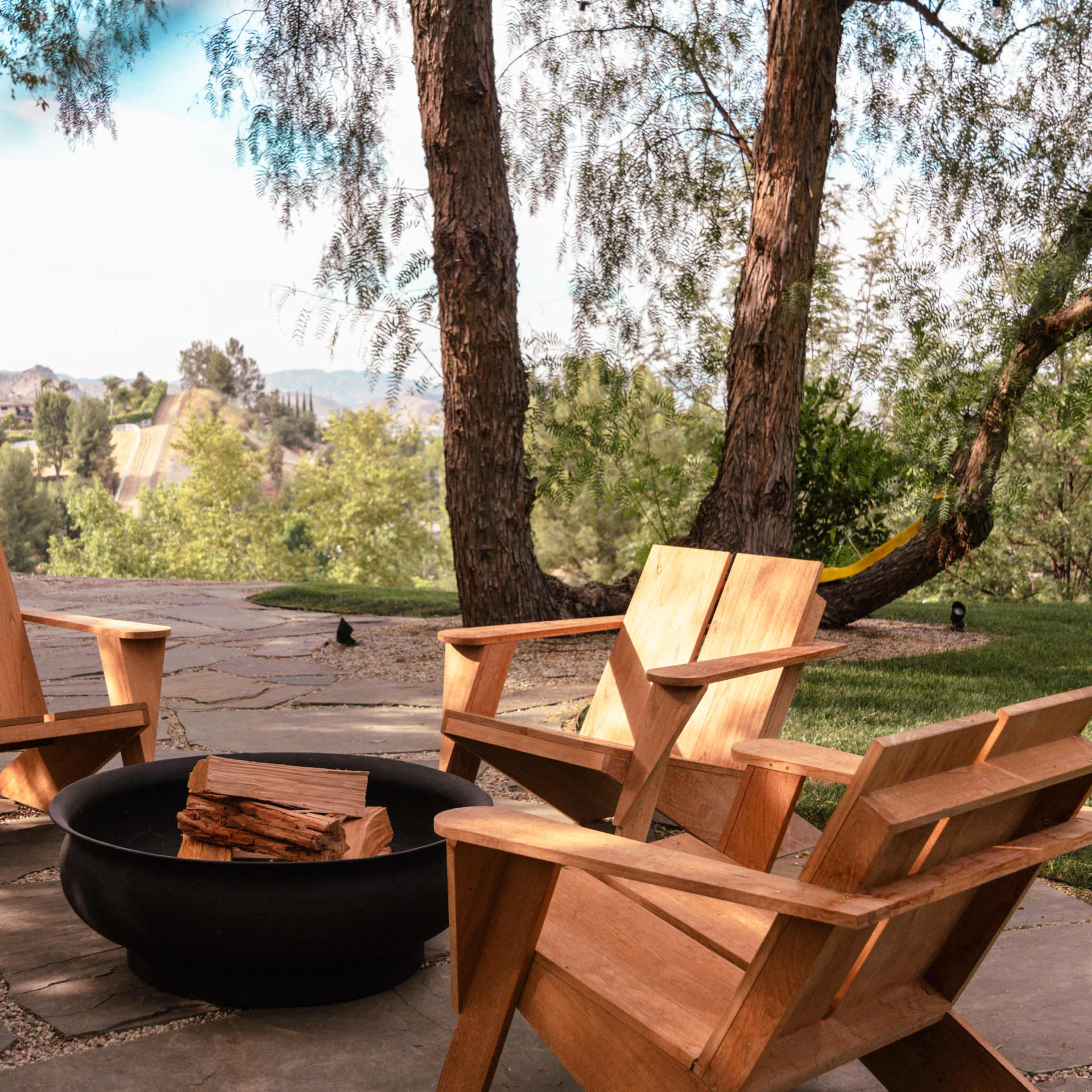 Four wooden chairs arranged around a fire pit on a stone patio, surrounded by trees and greenery.