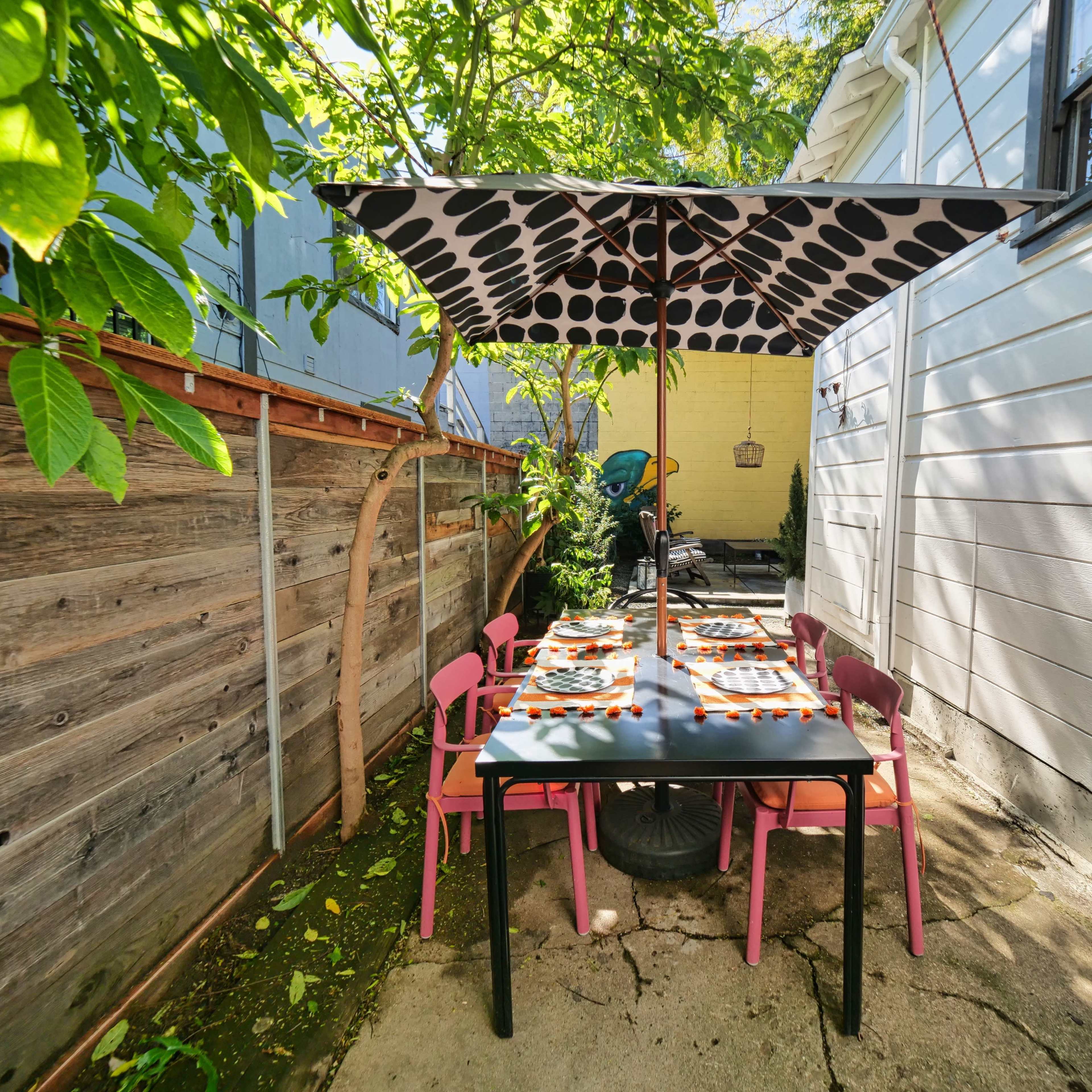 A dining table with colorful plates is set under a polka-dotted umbrella in a shaded outdoor space surrounded by greenery.