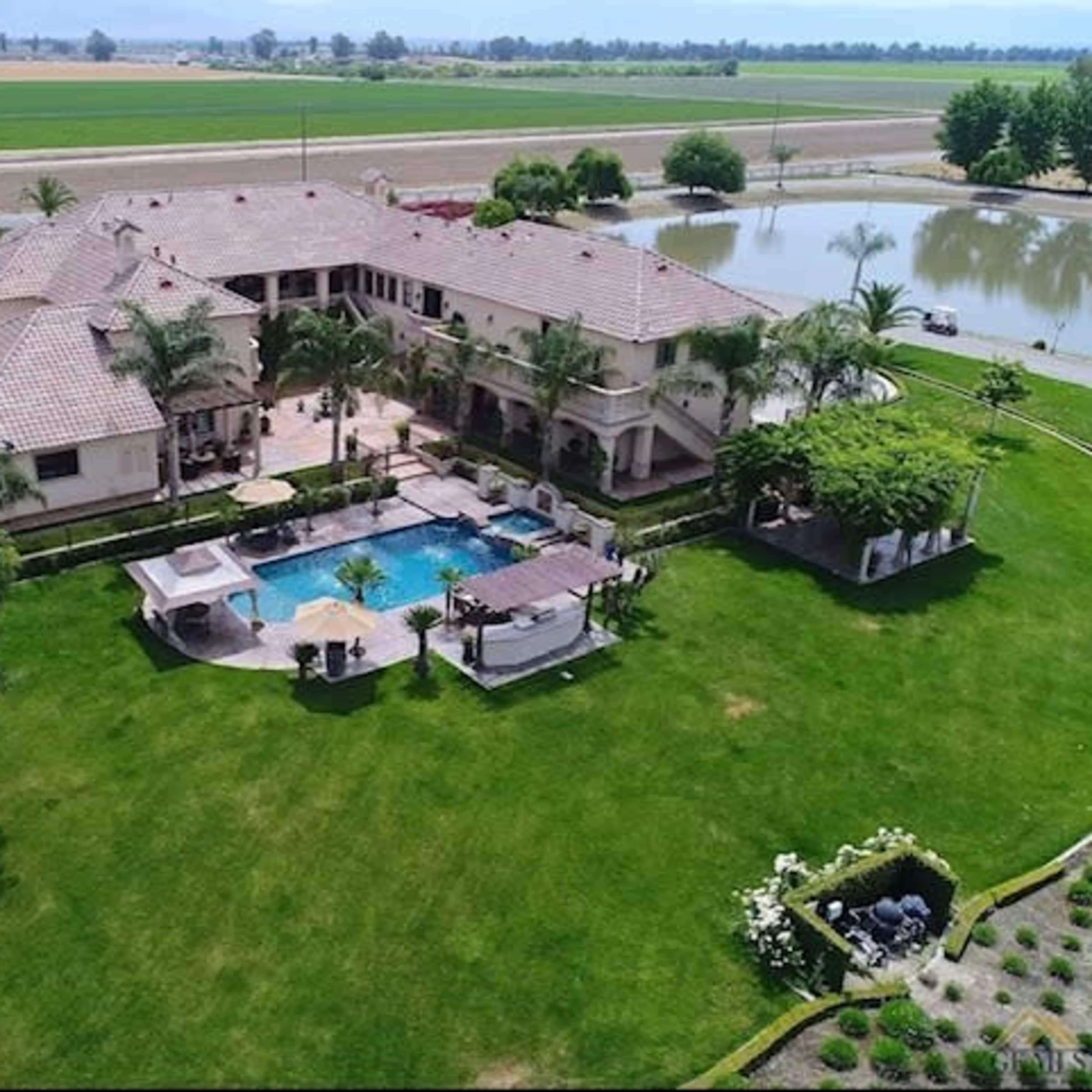 A large, multi-story house surrounded by green lawns, a swimming pool, and a pond, with fields in the background.