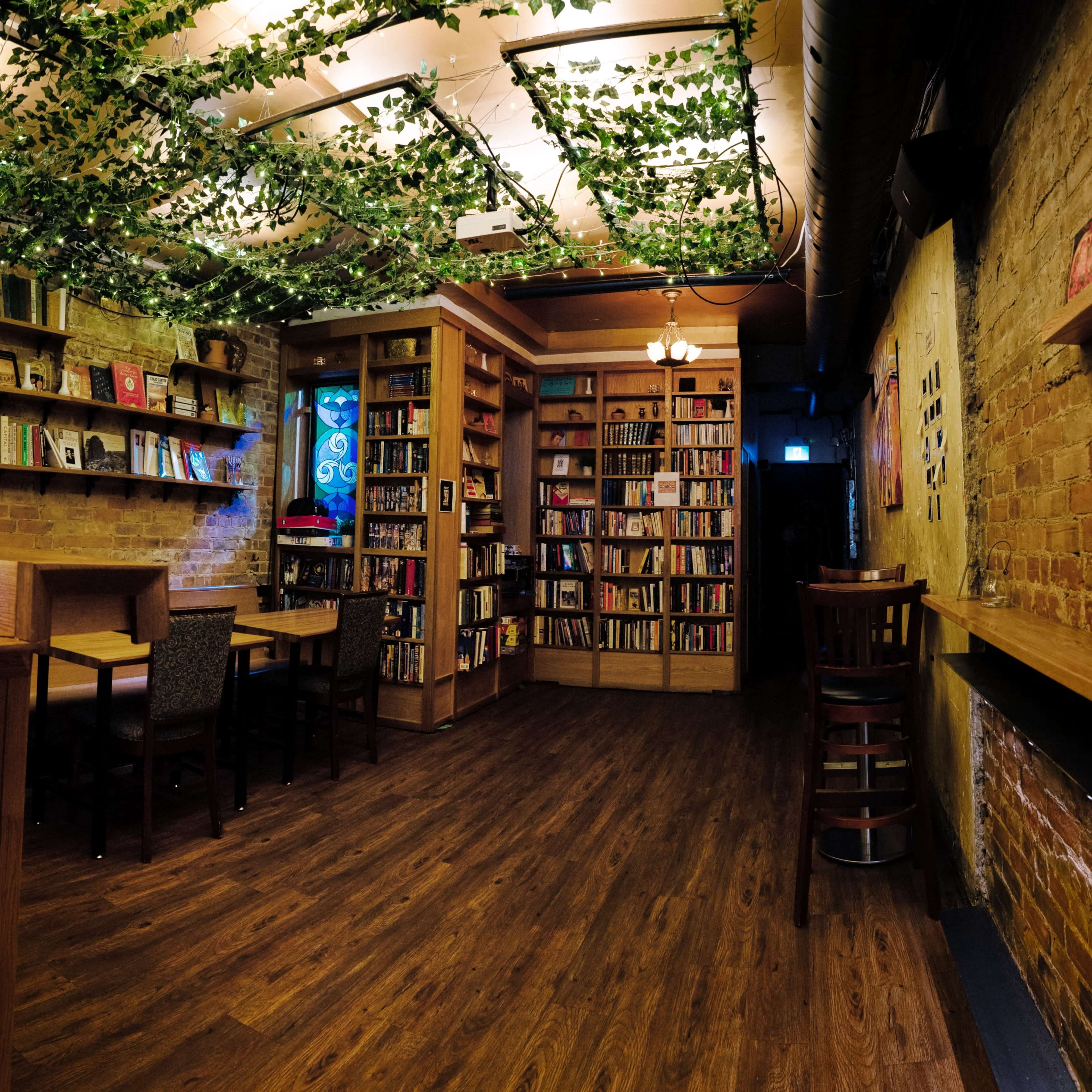 A cozy café features wooden tables, a bookshelf lined with books, and greenery hanging from the ceiling.