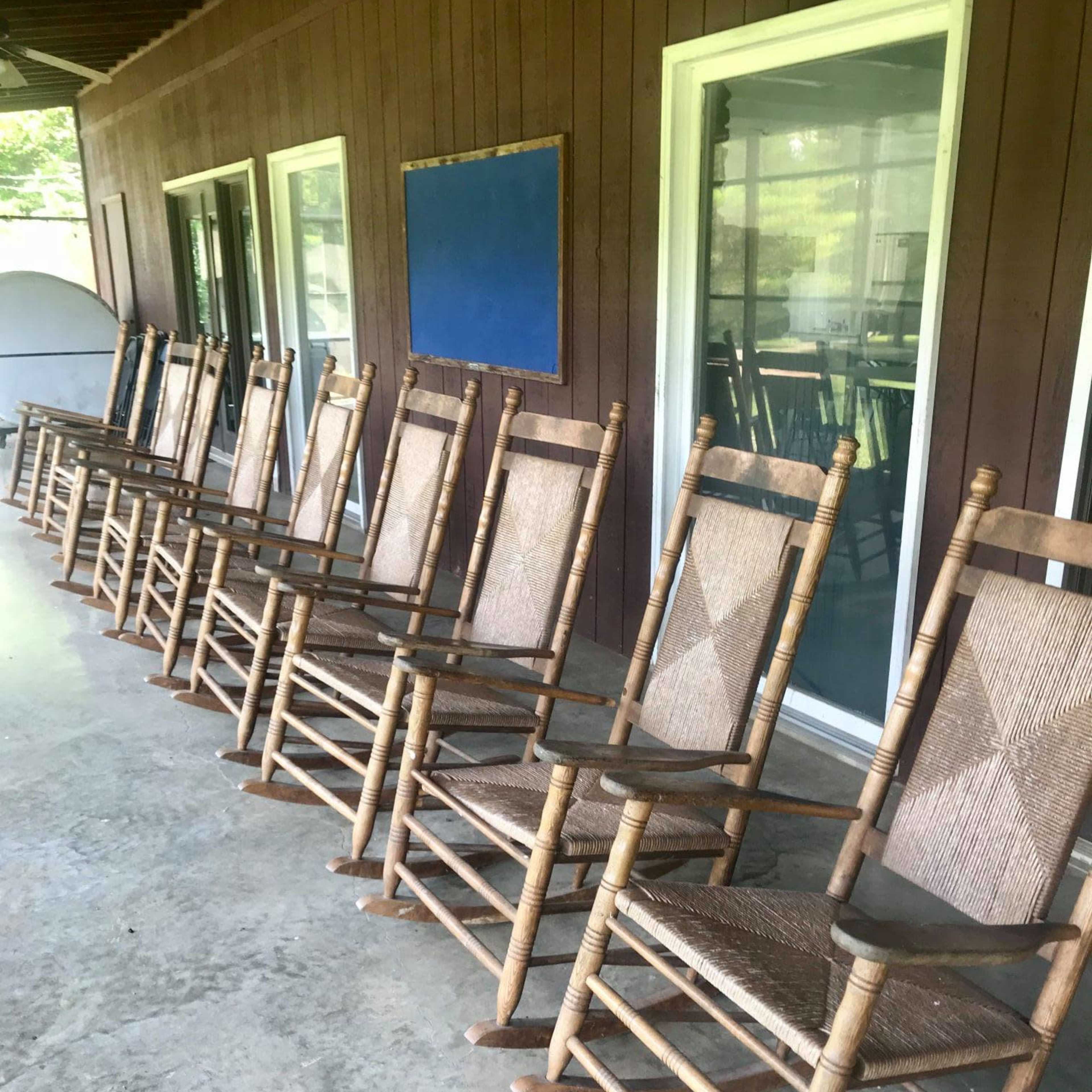 A row of wooden rocking chairs is lined up on a concrete patio beside a wooden wall with large windows.