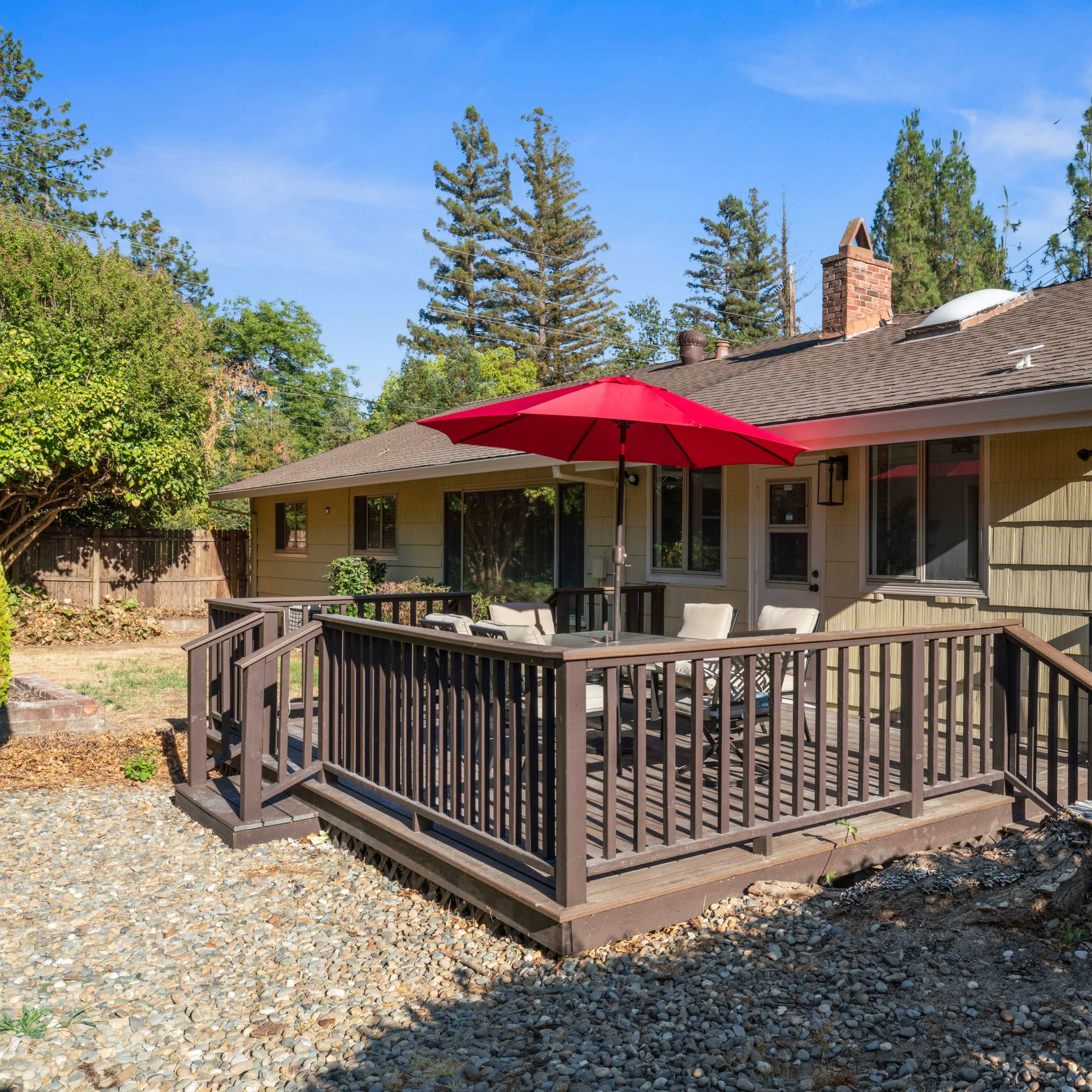 A yellow house with a deck featuring an umbrella stands surrounded by trees and gravel.