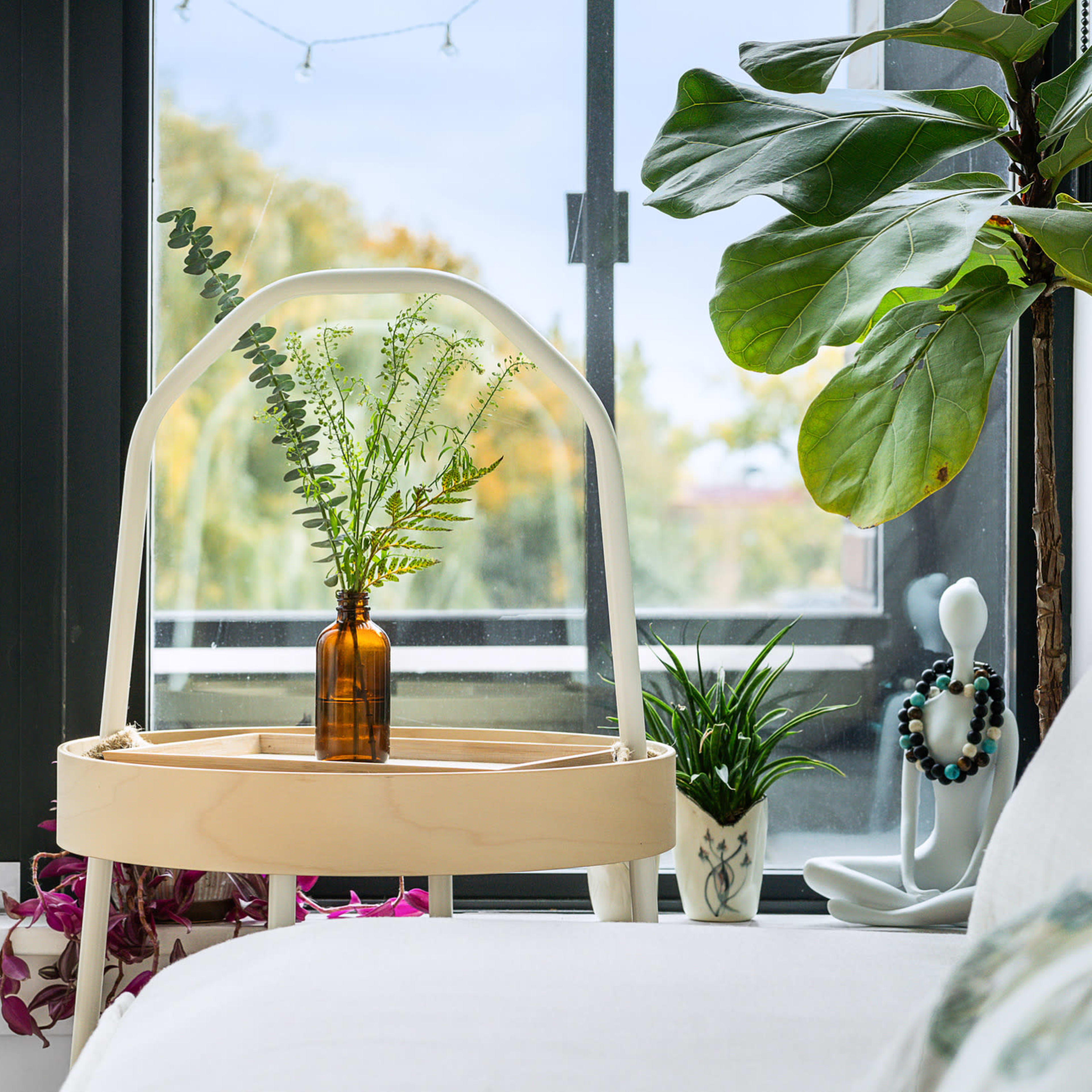 A minimalist side table with a glass cloche displays a bottle filled with greenery, positioned near a window alongside decorative plants and a sculpture.