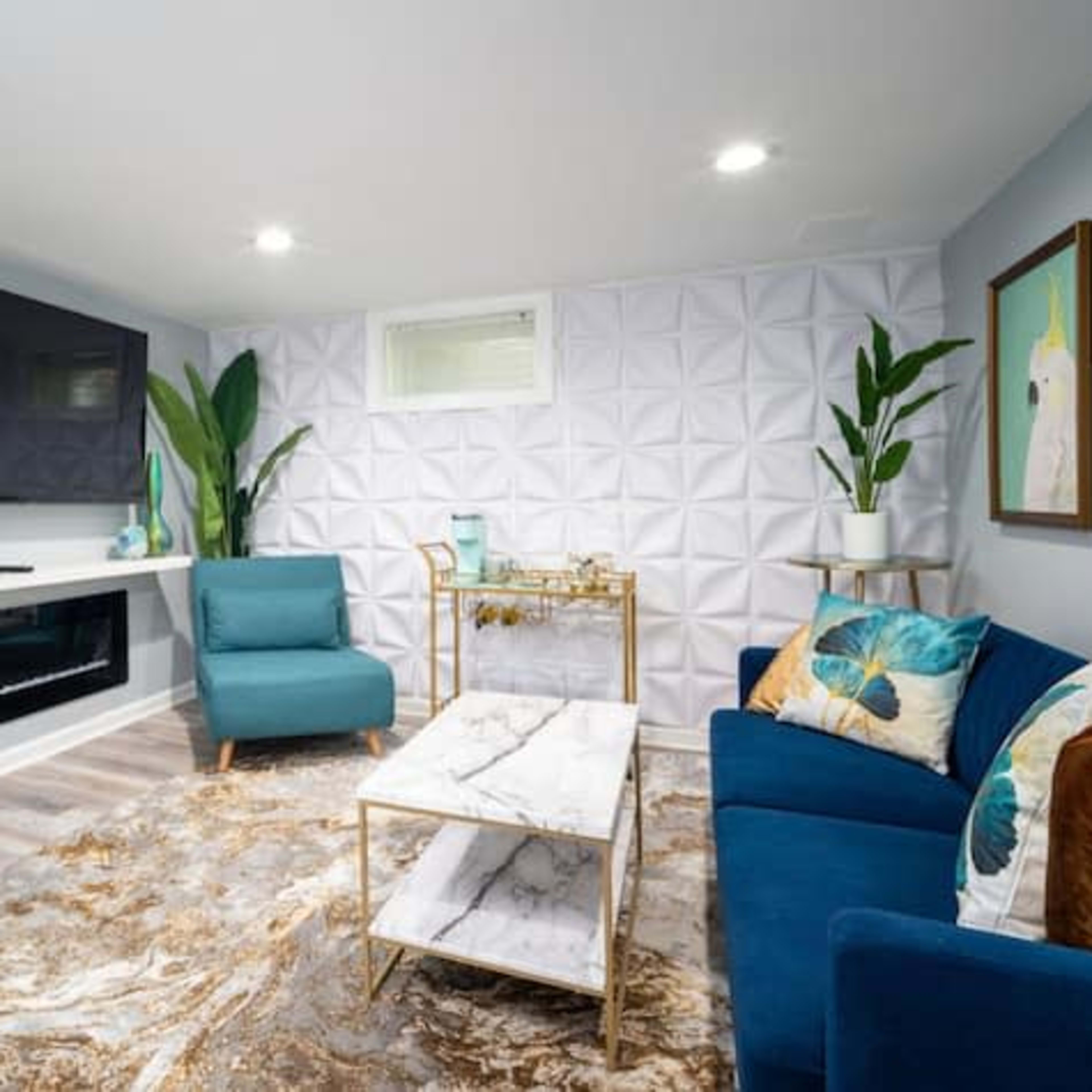 A modern living room with a large flat-screen TV mounted on the wall, a white textured backdrop, and blue seating arranged around a marble-topped coffee table.