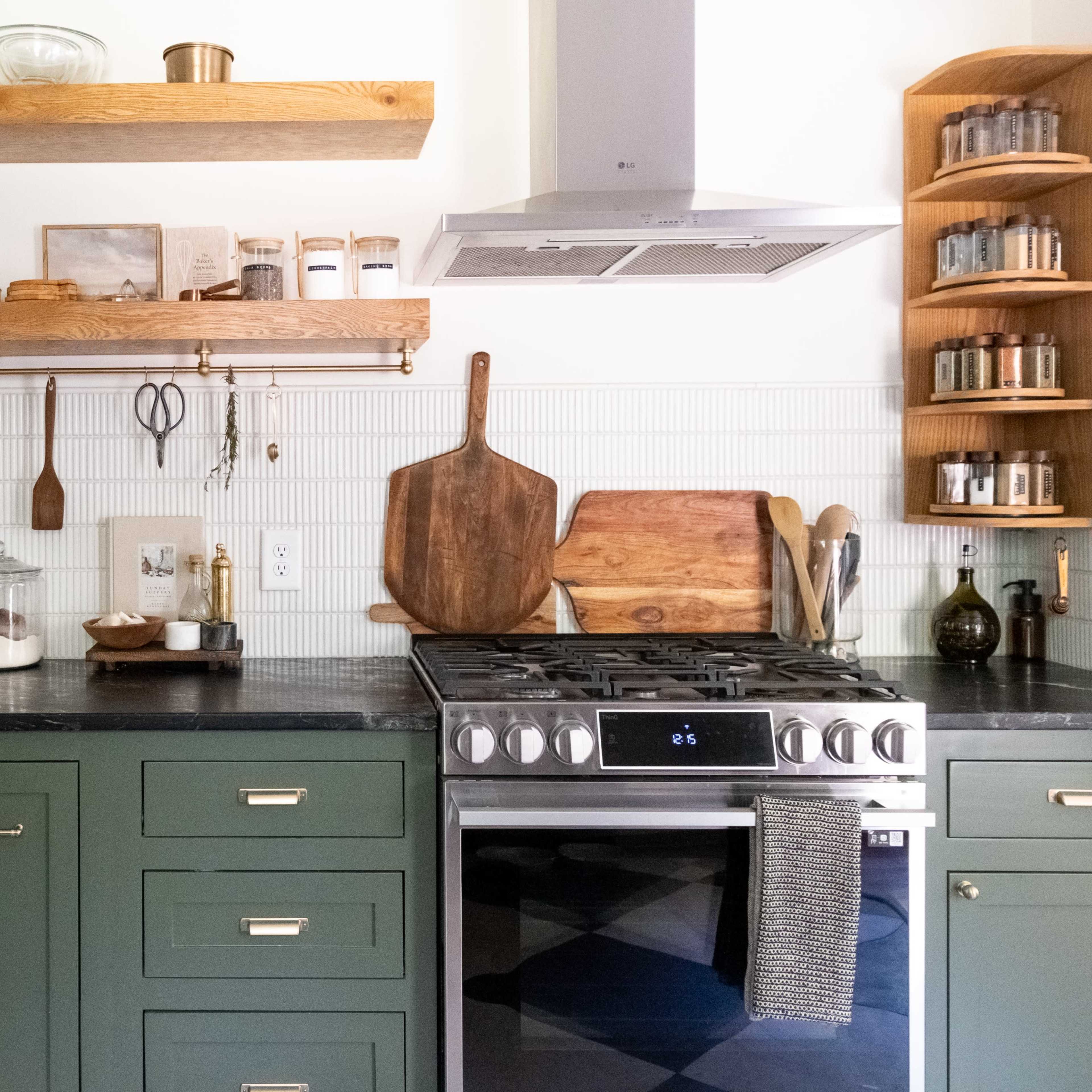 A modern kitchen features green cabinetry, wooden shelves with kitchen utensils, and a stainless steel stove against a white tiled backsplash.