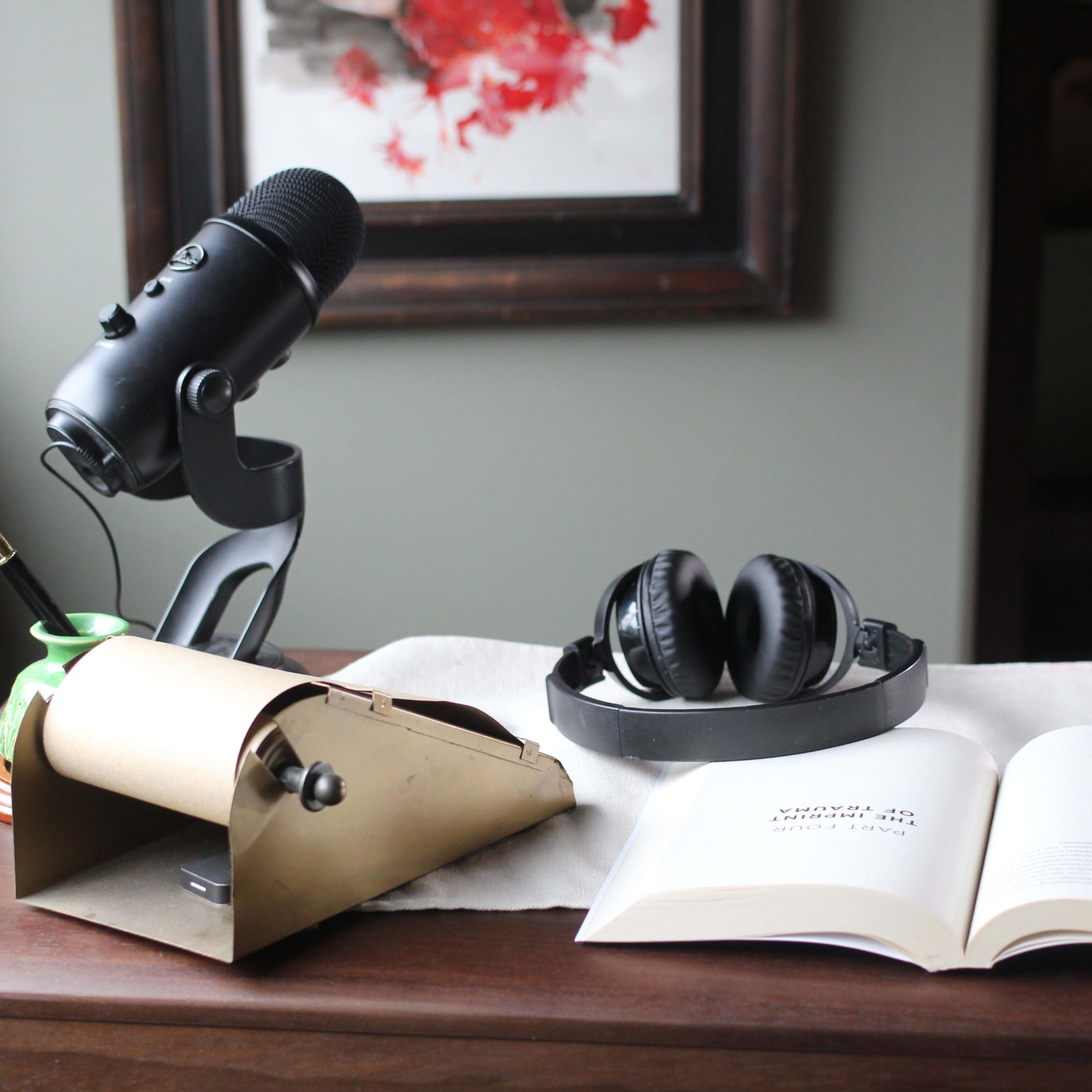 A black microphone and headphones sit on a wooden table next to an open book and a brass paper holder.