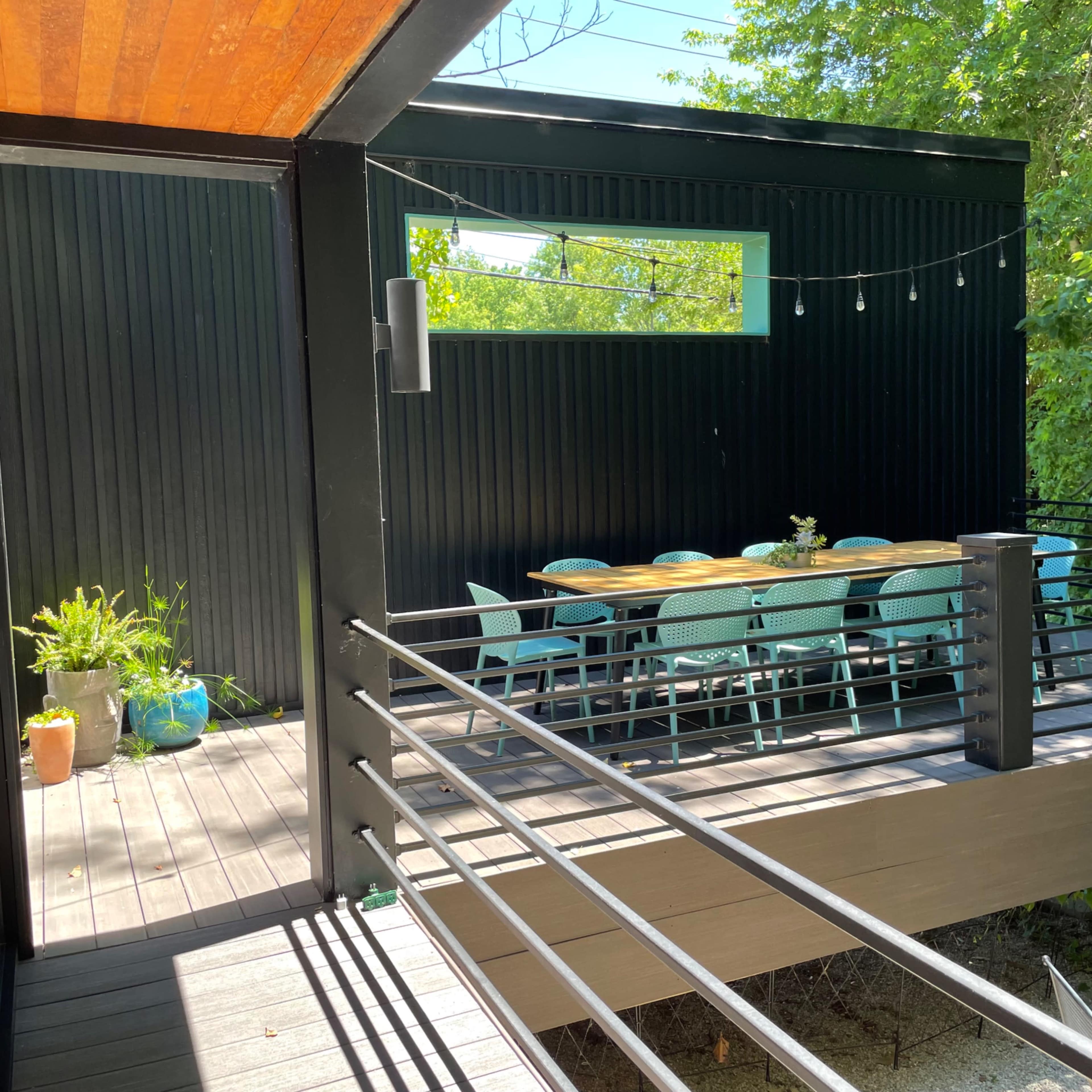 A wooden deck with a black-paneled wall features a dining table surrounded by turquoise chairs and potted plants.