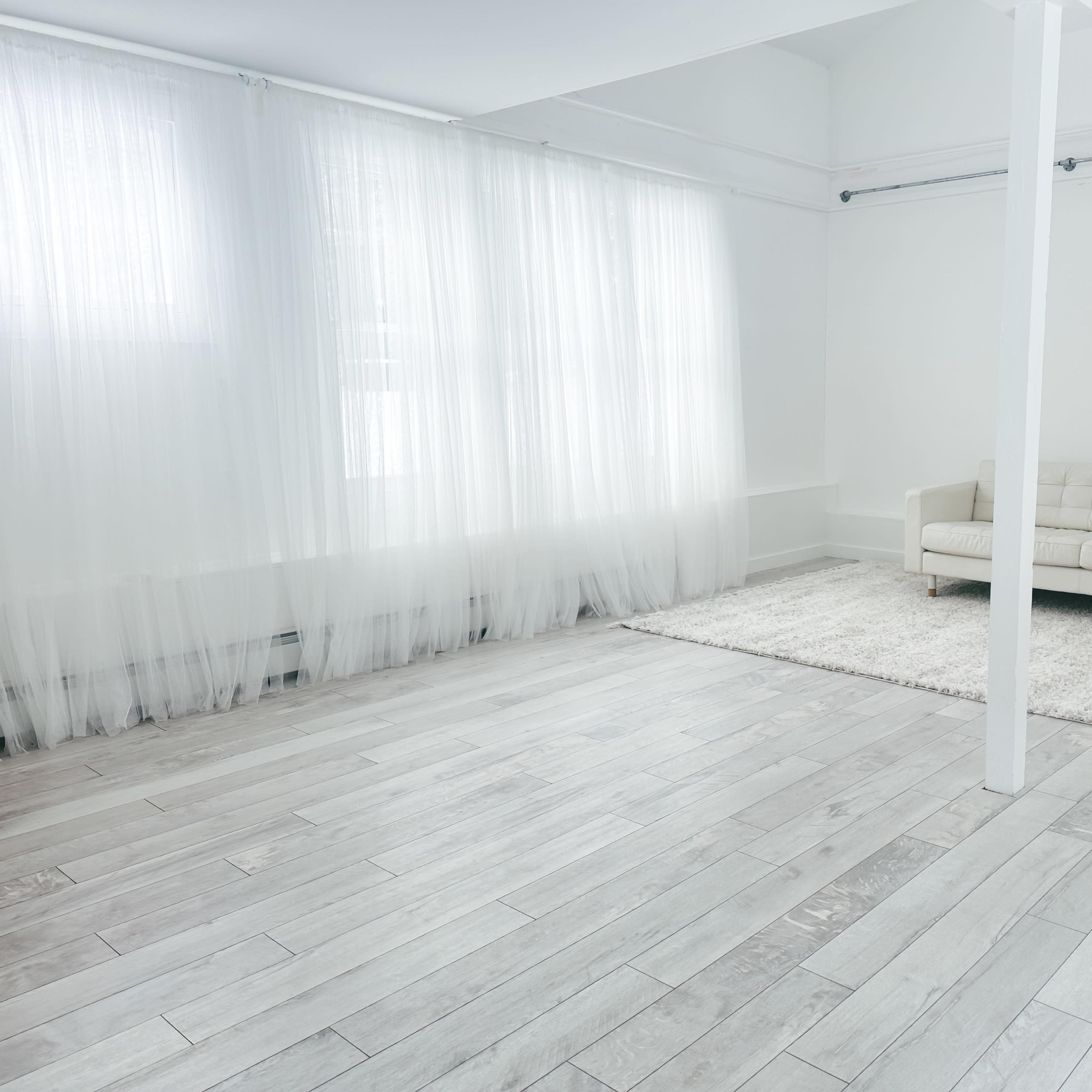 A bright, minimalist room with light-colored wooden flooring, a white sofa, and sheer curtains allowing natural light to filter in.