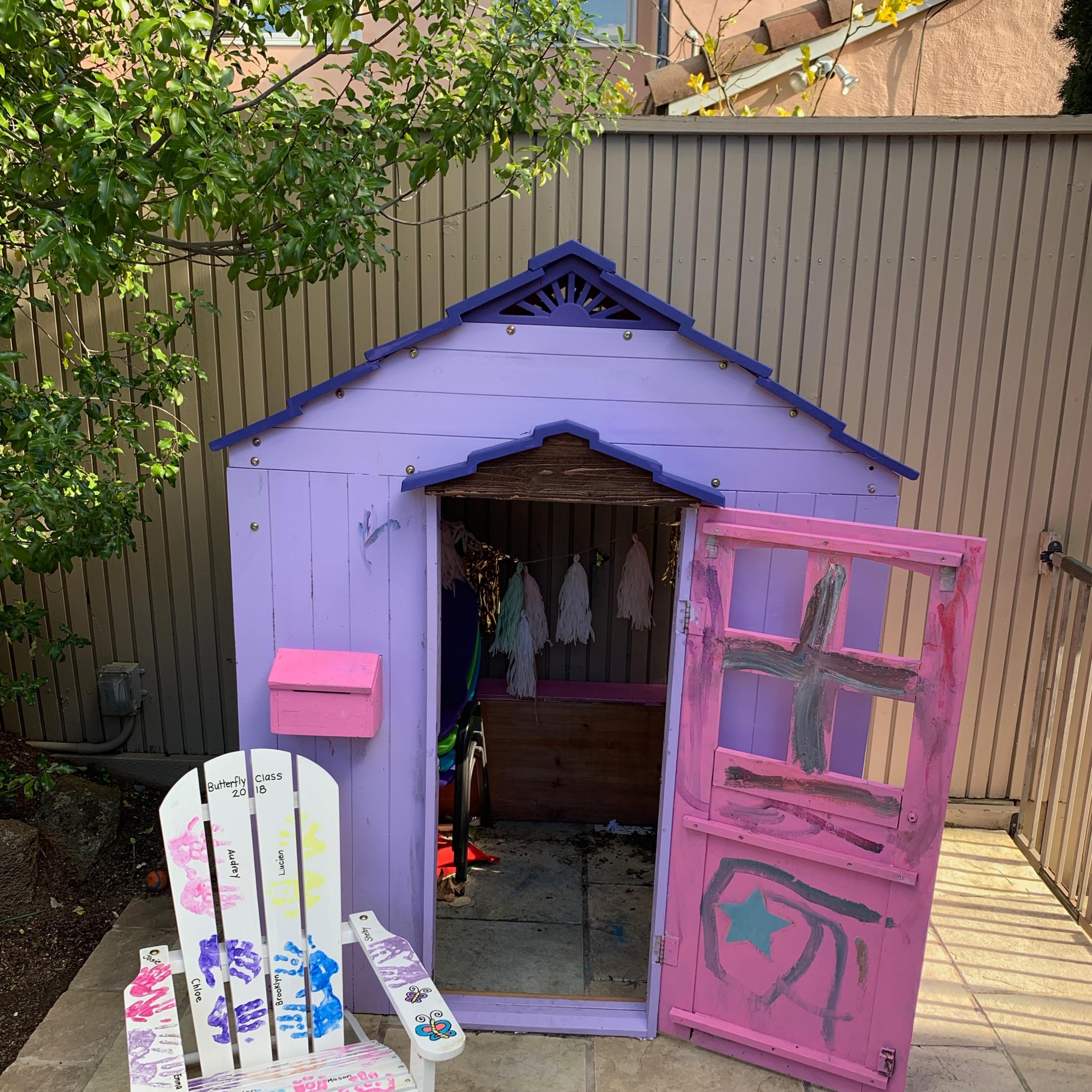 A small, colorful playhouse with a pink door and a white chair decorated with paint designs, set on a stone patio.